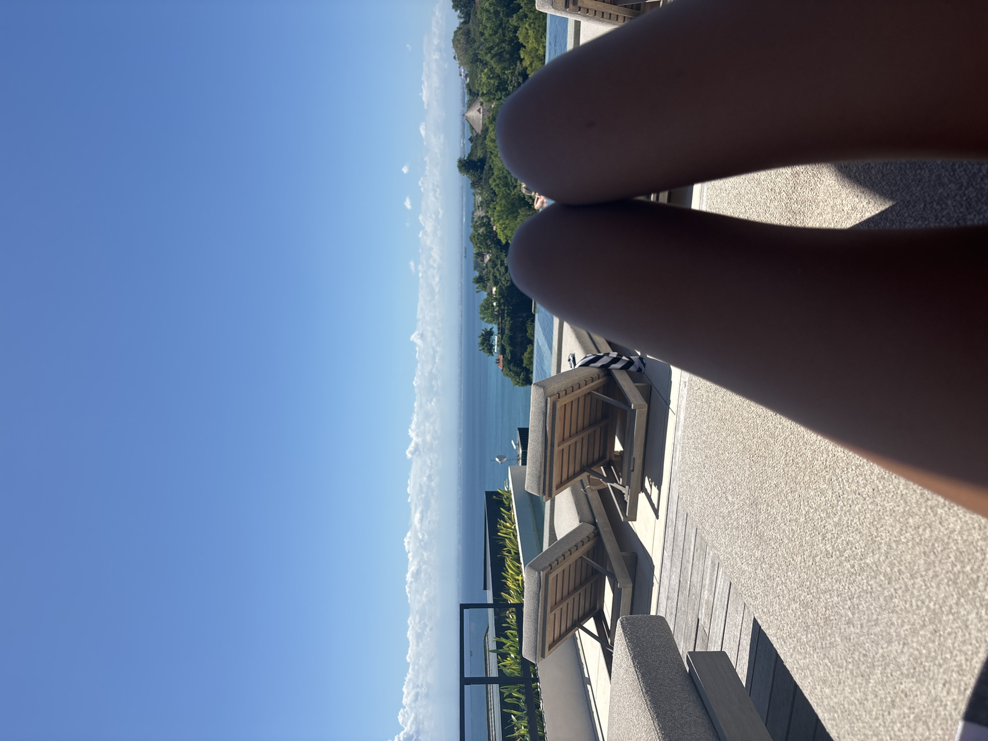 A first-person, upward-angled shot shows a person's legs resting on a textured, light-grey surface, likely a lounge chair. In the background, there's a vista of the ocean, a coastline densely covered in green vegetation, and a clear blue sky with some clouds near the horizon. Below the person's legs, lounge chairs are positioned near a glass barrier that overlooks the ocean. The location is identified as Kuta Selatan, Indonesia.