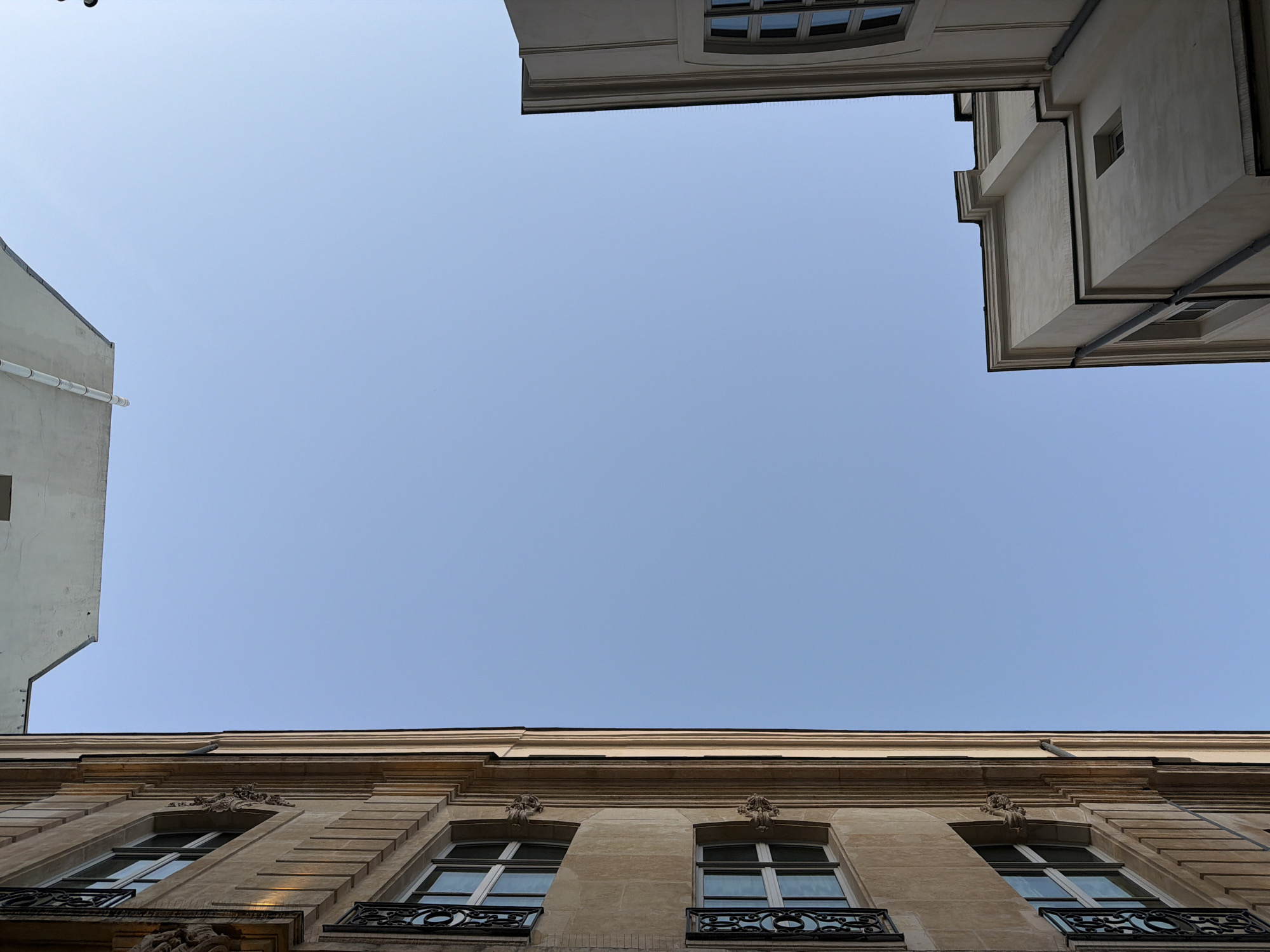 The image shows a view looking up at buildings in Paris, France. The buildings are of a classical style, with decorative stonework and windows. The sky is blue and mostly clear. The buildings are white and beige colors. There is a small bit of a drainpipe.