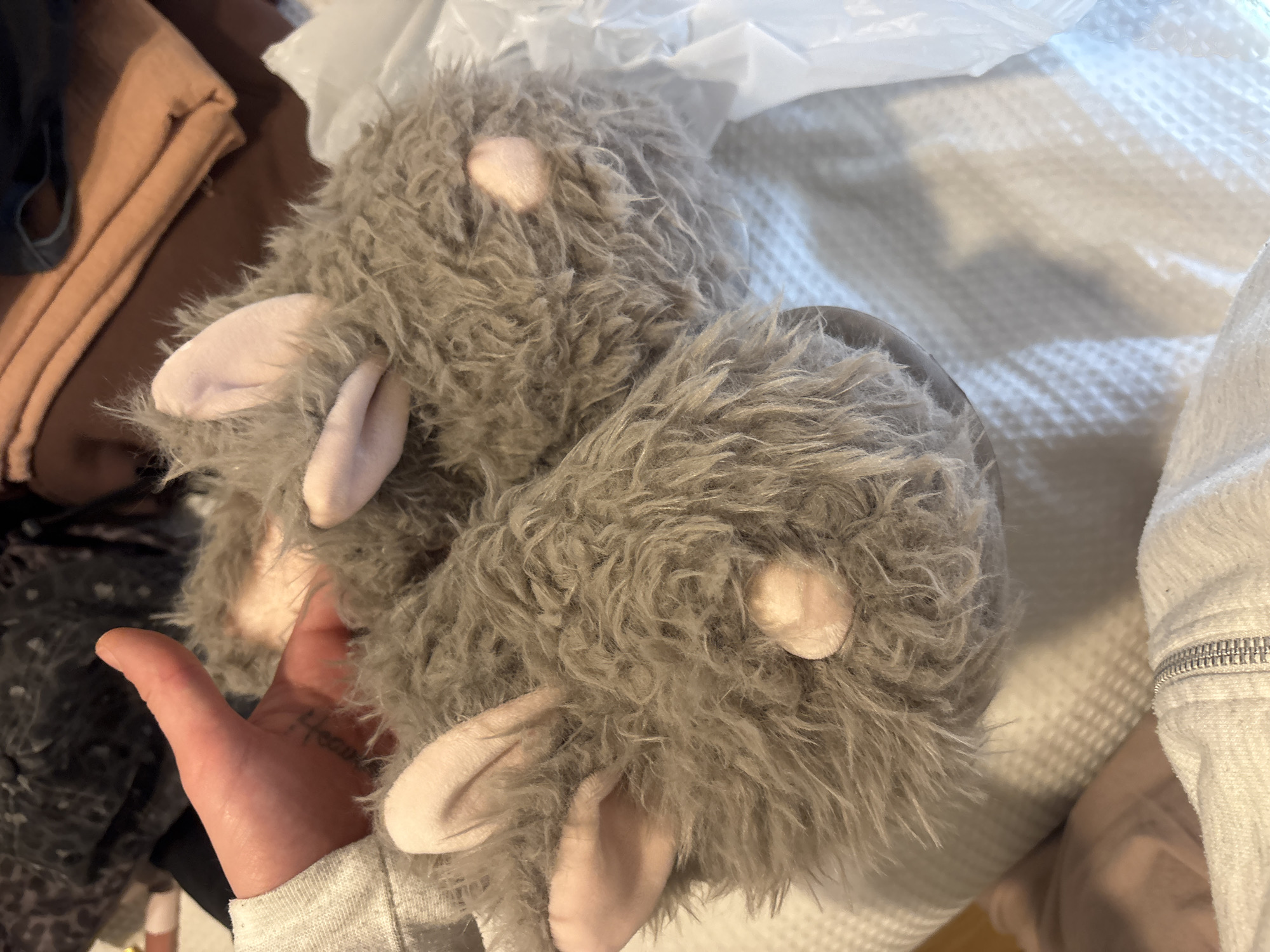 A pair of fluffy, grey animal-themed slippers dominates the center of the image. The slippers are characterized by their shaggy, long-pile grey fur, and each features two prominent, soft pink, rounded ears. A smaller, lighter pink circular detail on each slipper likely represents an eye or a nose, contributing to their whimsical, animal-like appearance, possibly resembling mice or a similar creature.
A human hand, visible in the lower-left, is holding one of the slippers. The hand displays a tattoo on the side of its thumb, which appears to spell "Heaven" in a stylized script font. The cuff of a grey, possibly knitted, long-sleeved garment is also visible on the hand.
The setting appears to be an indoor domestic space, likely a bedroom or dressing area. In the background, various clothing items are present: folded brown and dark blue fabrics to the left, and a white textured fabric, possibly a bedspread or blanket with a waffle-knit pattern, to the right. A clear, crinkled plastic bag or packaging material can be seen at the top of the frame. The lighting is soft and even, consistent with indoor conditions. No specific visual cues within the image directly indicate the provided location of Buenos Aires, Argentina.