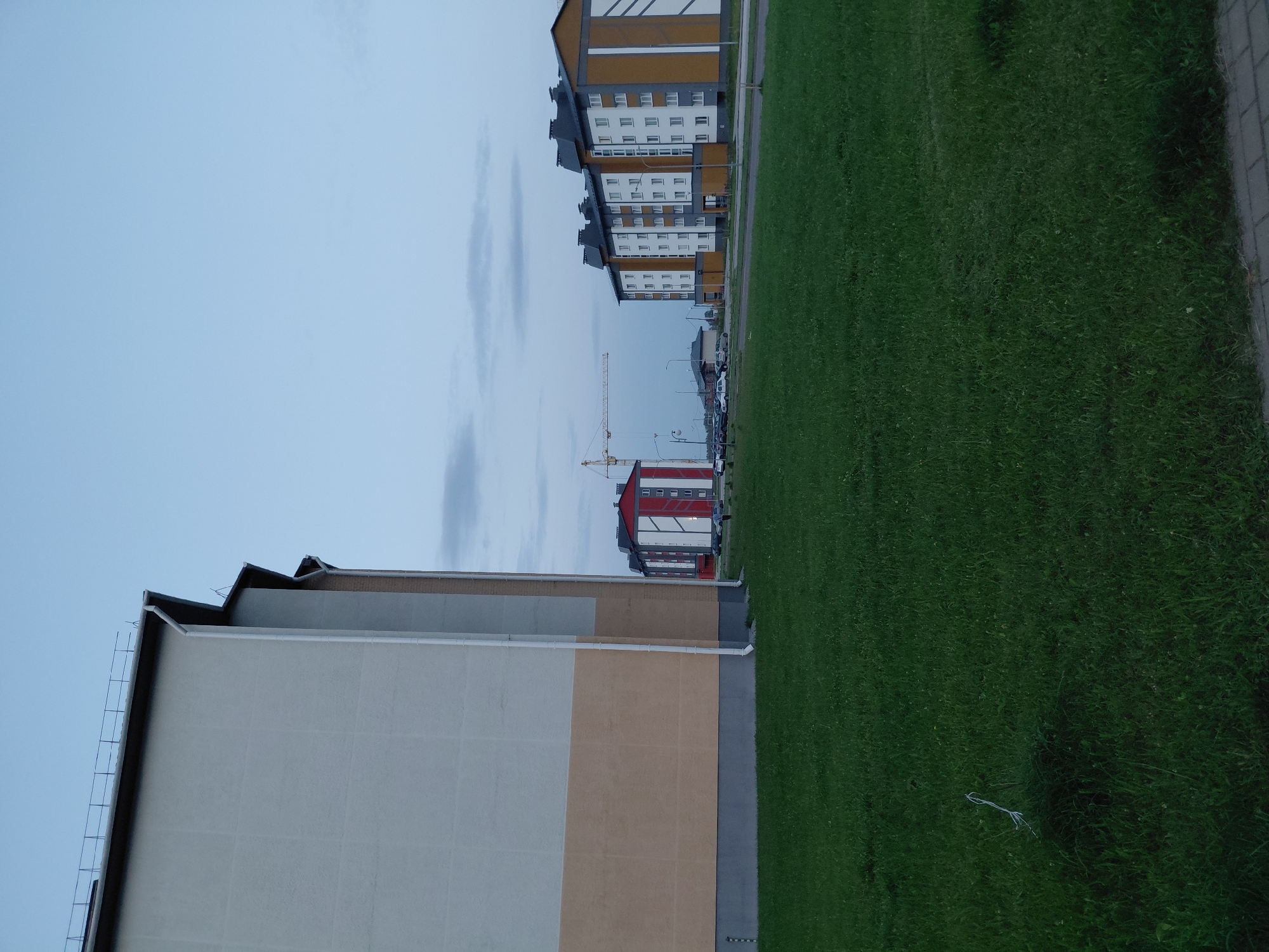 This image captures a scene in Smarhon, Belarus, likely during the early evening or dusk, given the subdued lighting and the pale blue sky with scattered clouds. In the foreground, a well-maintained green lawn dominates the lower half of the frame. A small piece of white string or cord is visible on the grass towards the left side.
Dominating the left side of the image is the side of a large, modern residential building. The building features a light beige upper section and a darker tan lower section, separated by a horizontal line. A white downspout runs vertically along the building's exterior. The building's roofline is visible at the top, with a dark railing along its edge.
Further into the background, other residential buildings are visible. To the right, a multi-story apartment building displays a modern design with alternating panels of white, orange, and gray. This building has a dark-colored roof with multiple dormer-like structures. To the left of this building, another apartment building is partially visible, distinguished by its red upper section with white vertical lines. A construction crane is visible behind these buildings, suggesting ongoing development in the area. Several cars are parked along the road in front of the apartment buildings. The overall environment appears to be a newly developed residential area within the city.