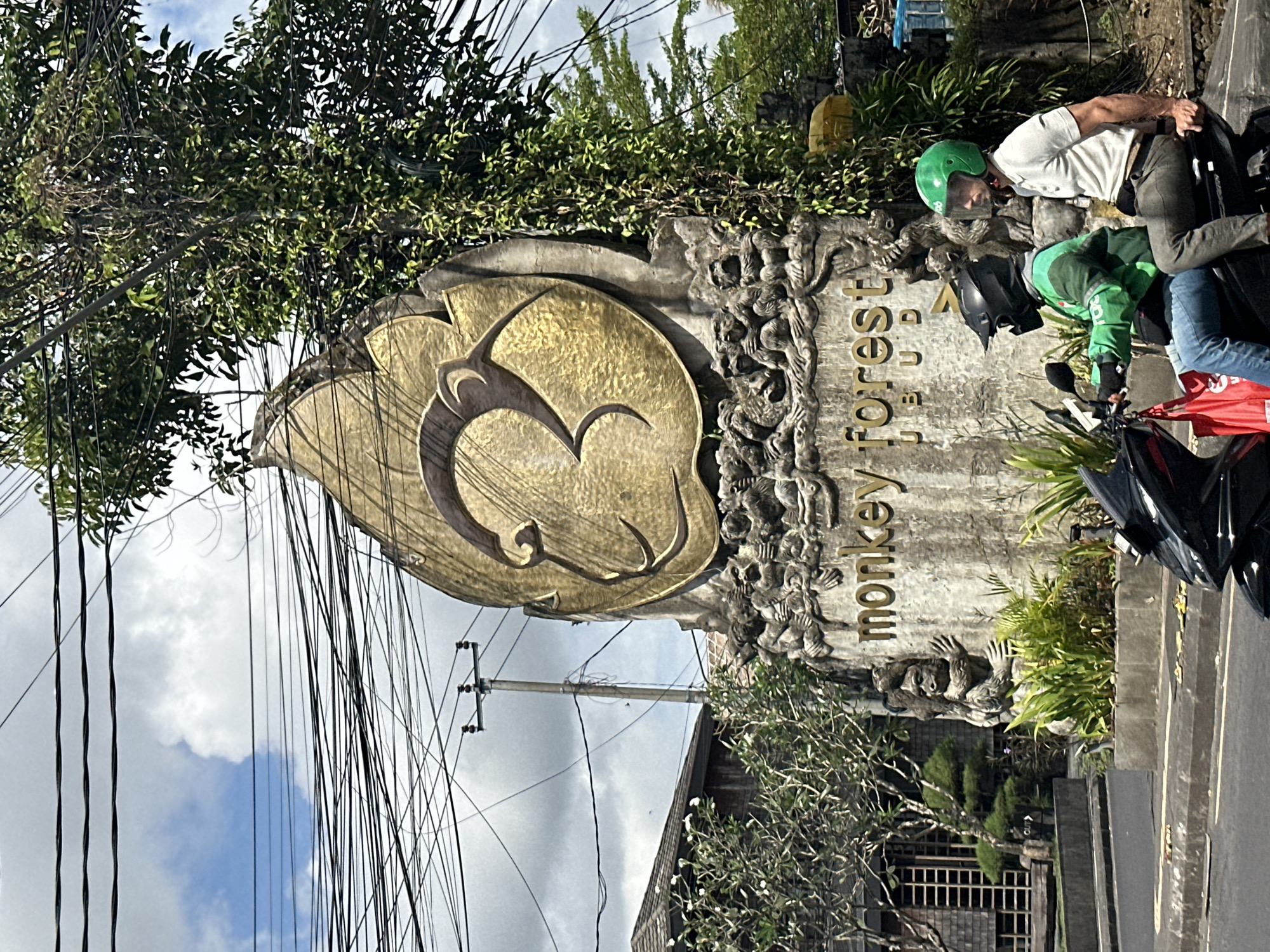 This image depicts the entrance sign for the Monkey Forest in Ubud District, Indonesia. The sign is a large, stone structure featuring a golden monkey face relief at the top, with intricate carvings of monkeys surrounding its base. Below the carvings, the words "Monkey Forest" are inscribed in gold lettering. The scene is framed by a lush, green tree on the right, and a building with wooden architecture on the left. Two individuals are riding a scooter in the foreground, one driving and the other as a passenger, both wearing helmets. Overhead, a dense network of electrical wires crisscrosses the sky, characteristic of the urban landscape of Ubud. The sky is partly cloudy, suggesting a bright day.