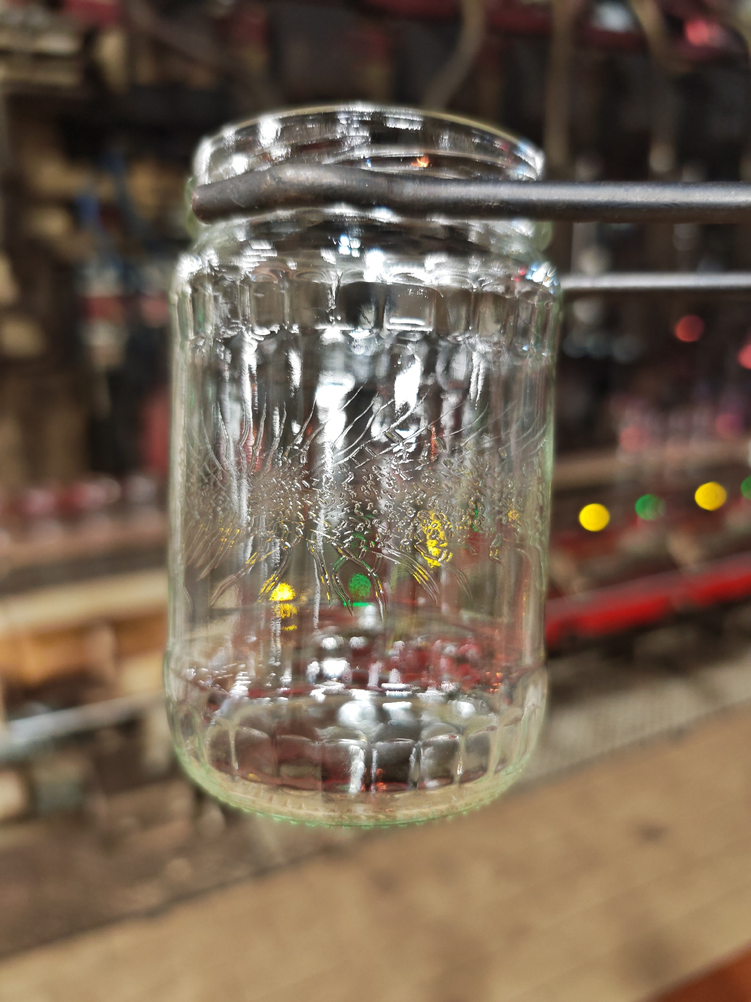 A glass jar with an embossed design is held by a metal clamp, likely in an industrial setting. The jar appears to contain a small amount of reddish liquid at the bottom. The background is blurry, but indicates an area with machinery and some colored lights. The exact location in the city of Pleven, Bulgaria is not discernible from the image. No people or interactions are visible.