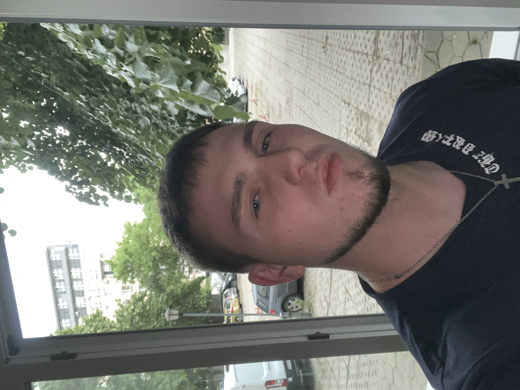 A man is positioned in front of a window, which looks out onto a street in Plovdiv, Bulgaria. Outside, trees, buildings, parked cars and a sidewalk are visible. The man is wearing a dark-colored t-shirt with white lettering and a silver chain with a cross pendant. The image is oriented vertically.