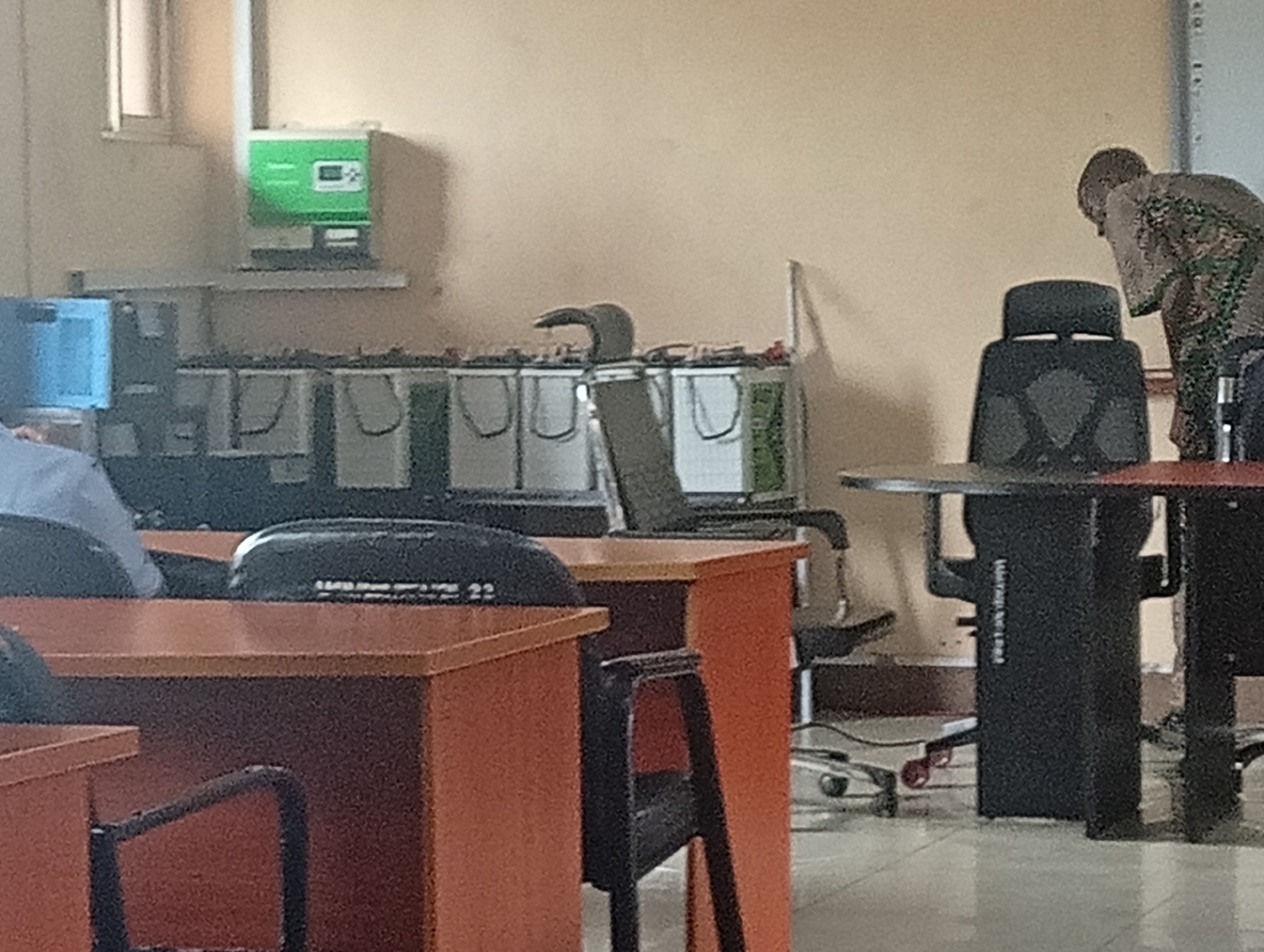 The image shows a classroom setting in what is likely an educational institution, possibly in Ijanikin, Nigeria.
**People:** There are at least two people present. One person is sitting at a desk in the foreground, visible from the side, wearing a light-colored shirt. Another person is standing by a table near a black office chair.
**Objects:** The foreground features orange-colored wooden desks and black chairs. In the background, there is what appears to be a power management system, likely a battery bank and possibly a green-colored inverter on the wall. Tables with a black base are also visible.
**Environment:** The classroom has off-white walls. The floor appears to be tiled. The lighting suggests an indoor setting.