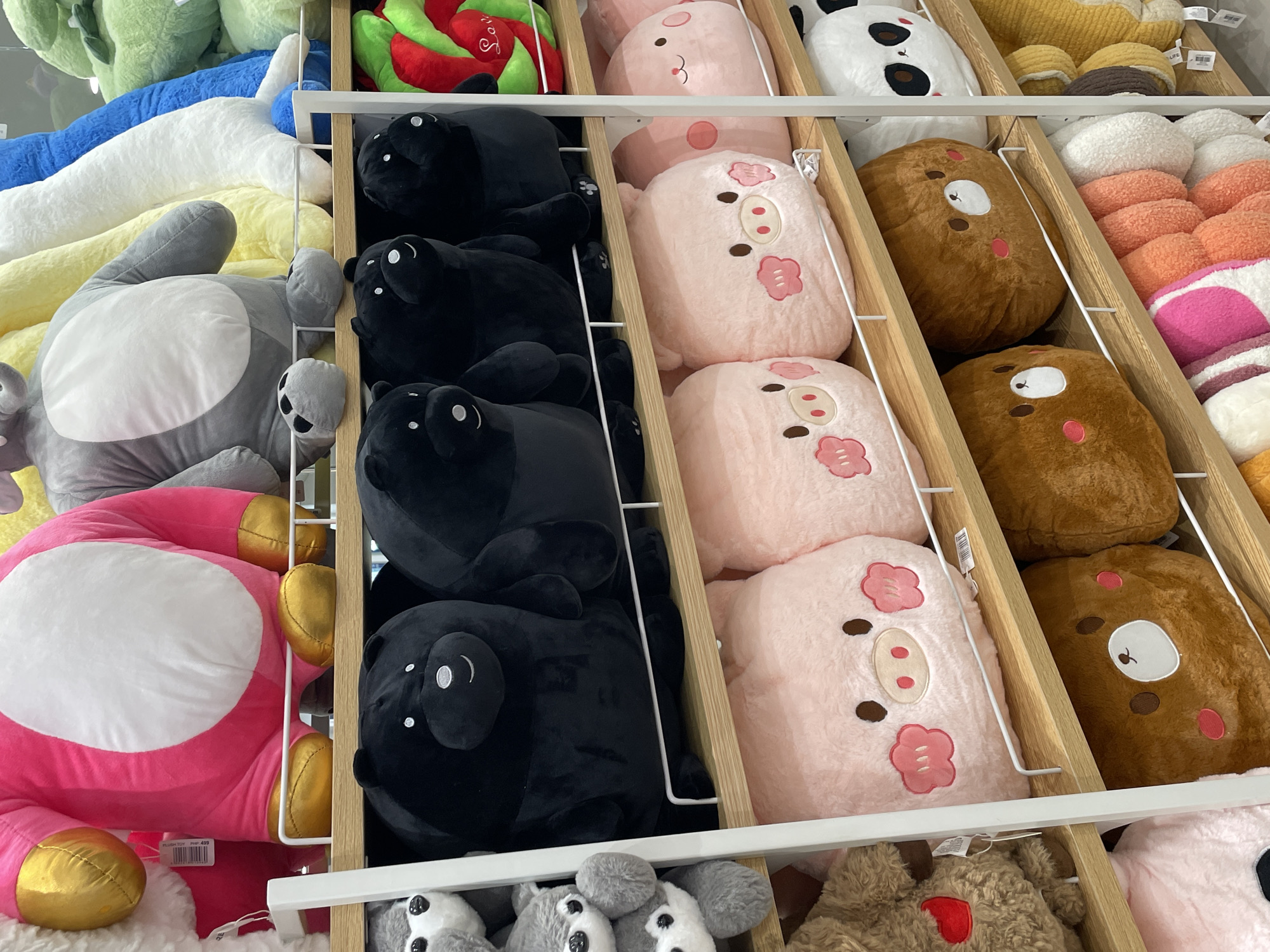 The image shows a retail display of plush toys on wooden shelves in a store. The toys are arranged in rows, with various animals like bears, pigs, and what appear to be seals or dogs. The toys are mostly large and brightly colored, with some featuring cartoon-like faces. In the background, other plush toys of different sizes and colors are visible. The overall environment suggests a toy store or a section of a department store. The image does not provide specific visual cues to identify the location as Davao City, Philippines, such as signage or distinct architectural features.