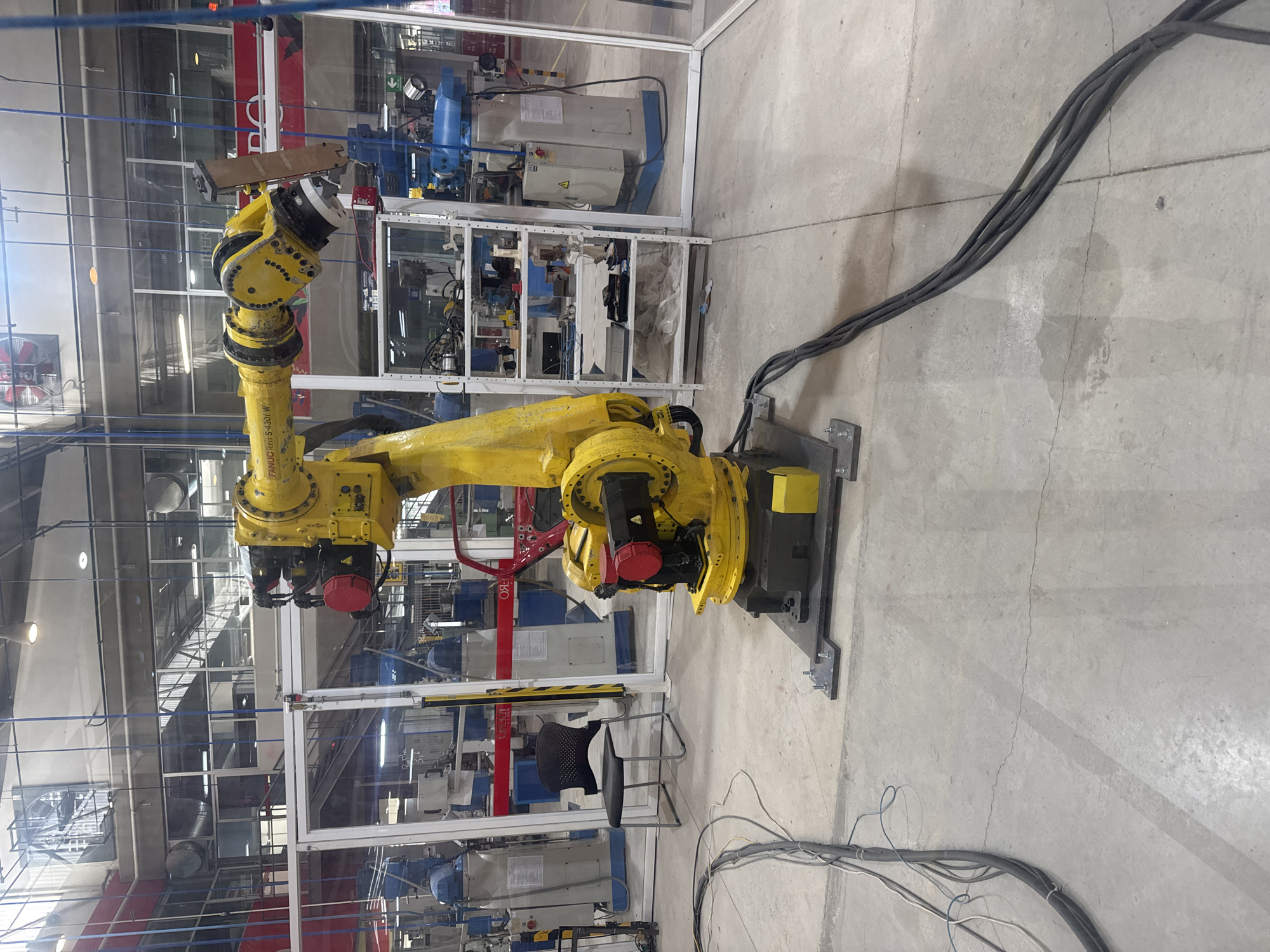 A yellow industrial robot arm is the central object in a factory setting in San Bernardino Tlaxcalancingo, Mexico. The robot, mounted on a gray metal base, is surrounded by clear plastic safety barriers, enclosing various pieces of industrial machinery. Black power cables extend from the base, running along the gray concrete floor. The environment appears to be a manufacturing facility with multiple pieces of equipment, overhead pipes, and scaffolding. A black chair is visible next to one of the barriers.