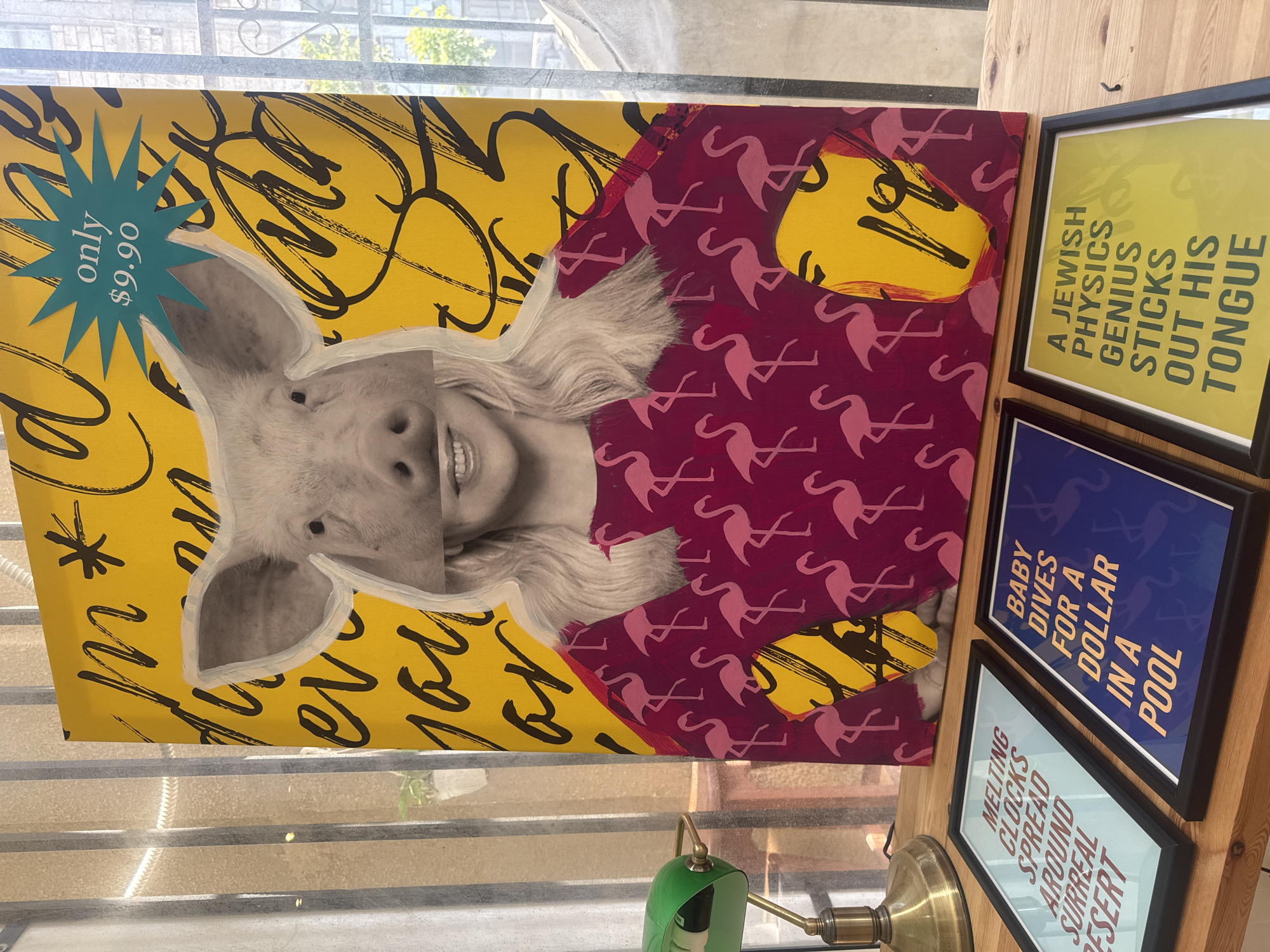 The image shows a collection of art pieces. One piece is a collage-style print of a pig's head with a woman's face, wearing a patterned shirt with flamingos, set against a yellow background with black scribbles. A blue starburst sticker reads "only $9.90". Adjacent to the print are three framed art prints stacked on a wooden surface. The top frame contains yellow text on a light green background that reads, "A JEWISH PHYSICS GENIUS STICKS OUT HIS TONGUE." The middle frame has blue text on a dark blue background that says, "BABY DIVES FOR A DOLLAR IN A POOL." The bottom frame has white text on a light blue background that reads, "MELTING CLOCKS SPREAD AROUND SURREAL DESERT." Part of a green and brass-colored lamp is visible in the foreground. The items appear to be indoors, possibly near a window. There is no verifiable information to confirm the location as Tel-Aviv, Israel.