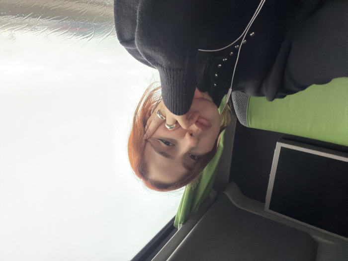 The scene depicts a young adult female seated inside what appears to be a bus or coach, located in Moineville, France. The female subject, with auburn hair, light complexion, and defined eyeliner, faces the camera directly, resting her right hand, adorned with two silver rings, against her jawline. Her lips are slightly pursed. She is wearing a dark, long-sleeved top featuring a high neck, silver stud embellishments along the chest, and a contrasting light-colored curved trim. White wired earphones are visible, with one earpiece in her left ear. To her left, a lime-green upholstered seat is visible, with a dark, unlit screen featuring a white bezel mounted on the wall behind it. To her right, a large window shows an overcast sky and a wet exterior, indicative of rain, with blurry red and white shapes, possibly distant vehicle lights, faintly discernible through the water-streaked glass. A folded green curtain is visible at the upper right edge of the window frame. The interior ceiling is dark gray or black.