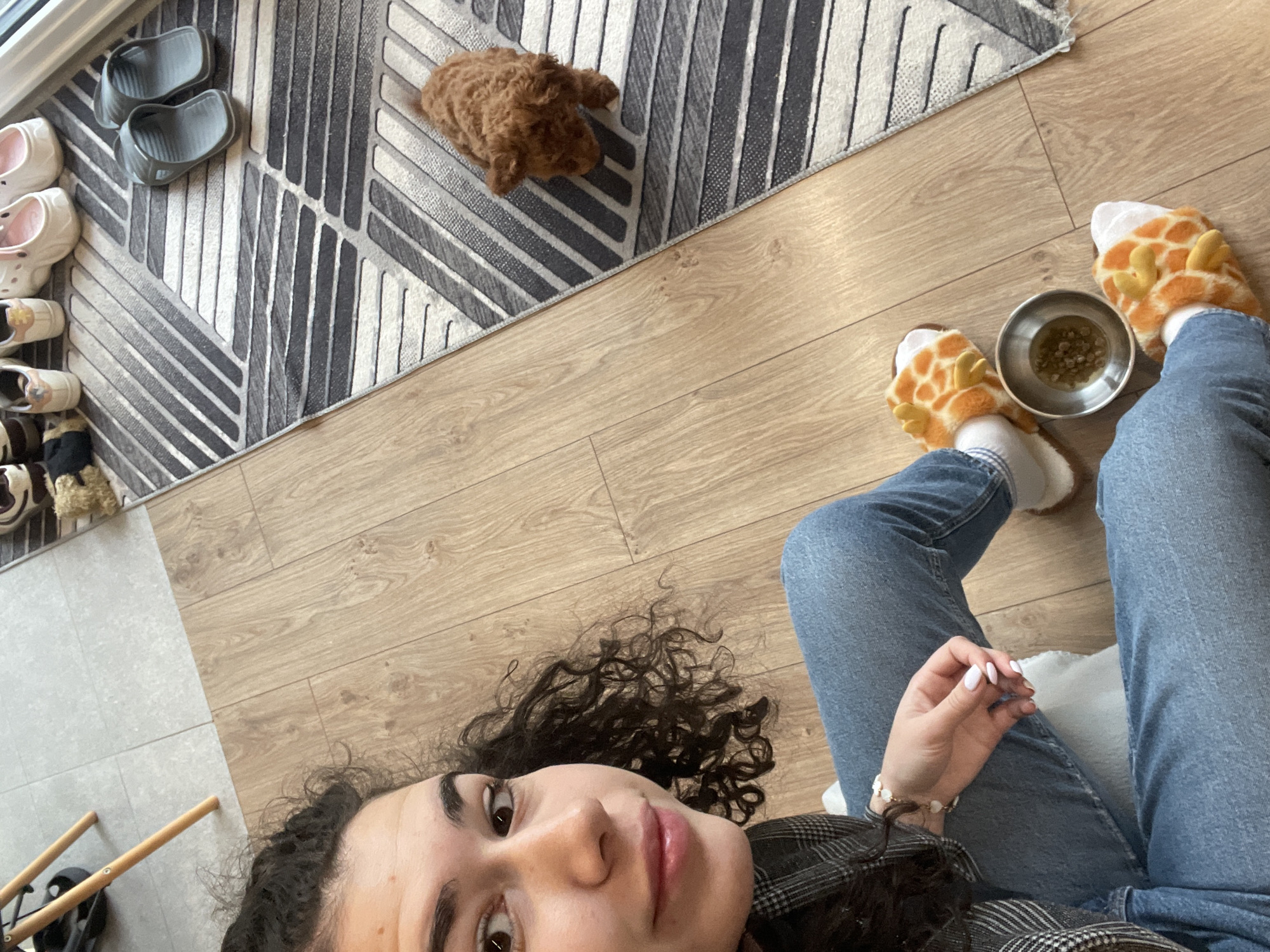 A woman with curly hair is taking a selfie, seated on a white pillow in what appears to be an apartment. She is wearing blue jeans and giraffe-themed slippers. Next to her is a metallic bowl filled with what seems to be animal food. A brown, poodle-type dog is lying on a gray and white patterned rug in the background. There are other pairs of shoes by the rug. The location is likely a residential apartment, but the city of Sofia, Bulgaria is not discernible from the image. The woman is holding her hand up near her face.