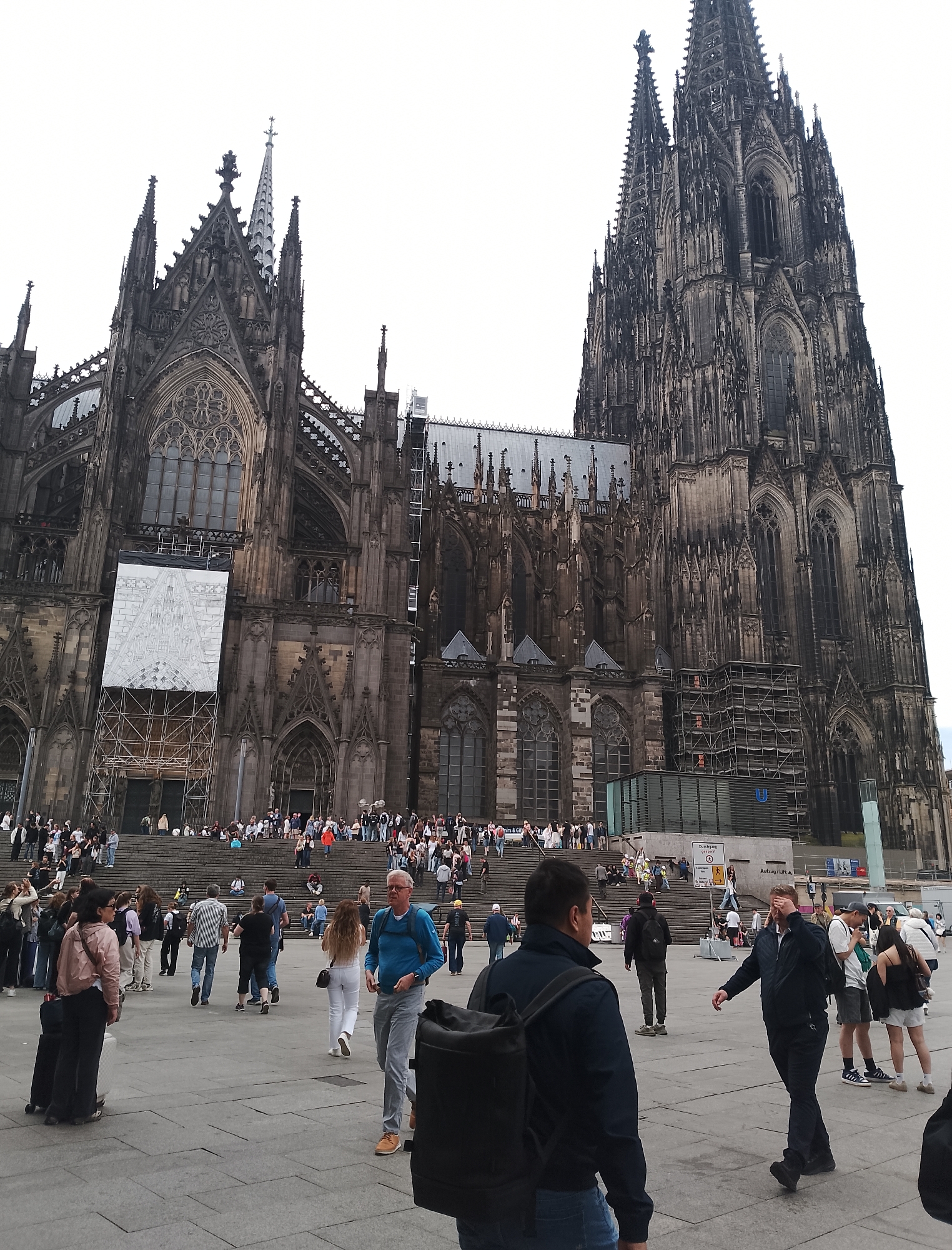 This is a wide-angle, eye-level, outdoor, daytime shot of a large Gothic cathedral. The cathedral features intricate stonework, pointed arches, and tall spires, with scaffolding visible on the exterior. A large banner hangs on the front facade. In the foreground and midground, numerous people are walking, standing, and sitting on wide stone steps leading up to the cathedral. The ground in front of the steps is paved. A few individuals are also visible in the immediate foreground. The sky is overcast.