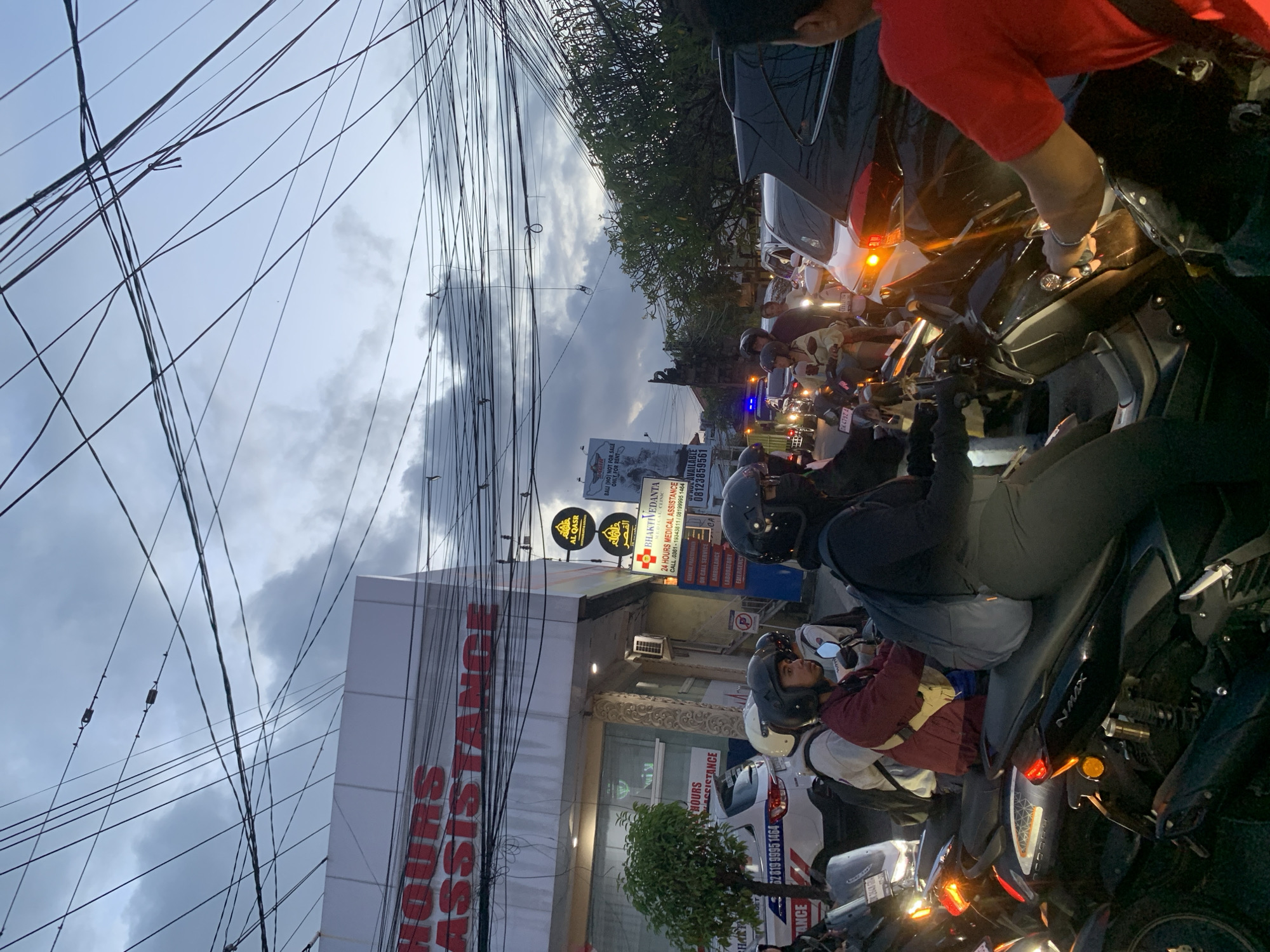 The image depicts a street scene in North Kuta, Indonesia. A crowd of people on motorcycles are clustered on the road. In the background is a building with signage for "24 Hours Medical Assistance" and names "AL QASR". Utility wires are visible overhead against a cloudy sky. A sign above the building advertises medical services, and a billboard is visible further down the road. A white car is parked near the medical building. Several people are wearing helmets.