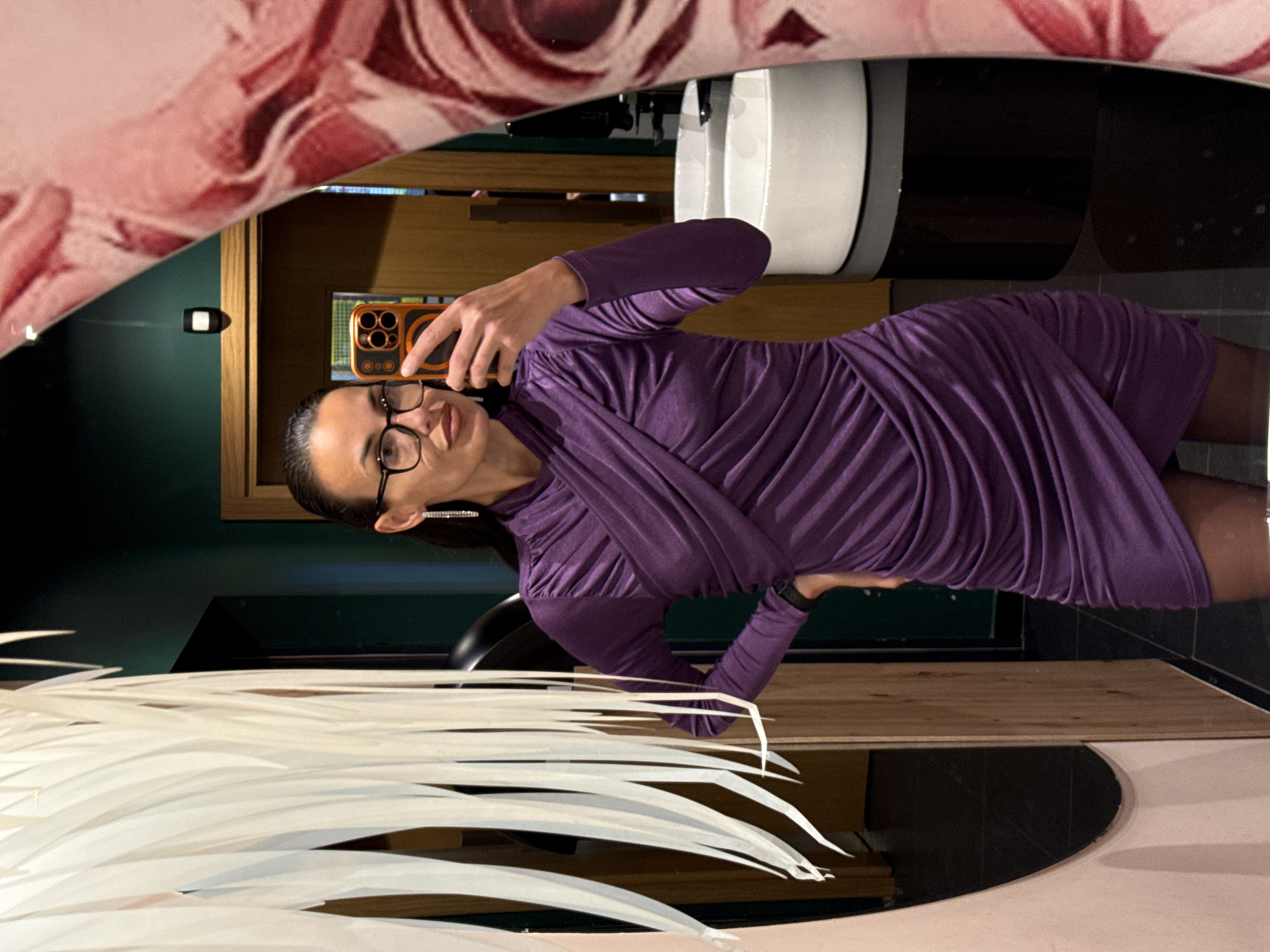 A woman is taking a selfie in a mirror, showing a bathroom interior. The woman is wearing a purple dress and glasses, holding a phone with an orange case. The mirror has a floral design on its upper left edge and a light pink rim. In the background, there is a white sink, dark green walls, wooden cabinets, and dark floor tiles. In the lower left corner of the mirror, there are decorative white feather-like objects. There is no evidence in the image to suggest the location is in the city of Pleven, Bulgaria.