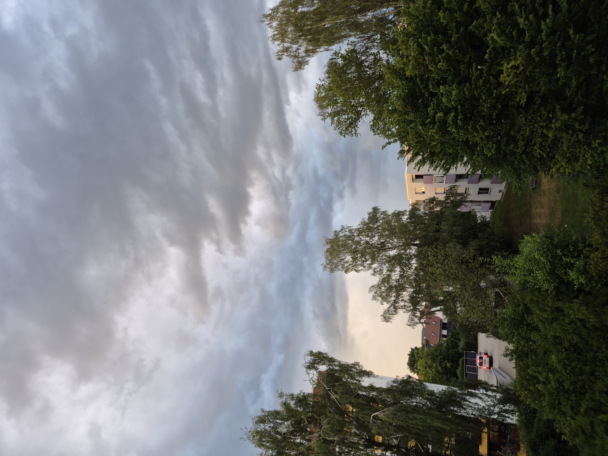 The image presents an elevated view of a residential area in Ergolding, Germany, under a heavily overcast sky. The upper two-thirds of the frame are dominated by dense, multi-layered grey clouds, exhibiting various shades from light to dark, with a subtle warmer, lighter hue visible in the lower-left sky quadrant. The lower third features extensive green vegetation, including several tall deciduous trees with light green foliage (appearing to be birches) framing the left and right sides and prominent in the mid-ground. A large, densely leafed, darker green tree occupies the mid-right. Below, a thick layer of green bushes and undergrowth fills the immediate foreground.
Architectural elements include:
1. A multi-story building, light-colored with yellow accents, partially visible on the far left, obscured by trees.
2. In the mid-ground to the left, a cluster of single-family houses with reddish-brown pitched roofs is visible, one featuring solar panels.
3. A paved driveway or parking area is located among these houses, where a white passenger car is parked, facing away from the viewer, with its red taillights illuminated.
4. To the right, another multi-story apartment building, predominantly white or light grey, features distinct purple/lavender balcony facades.
5. A patch of mown grass with a single wooden bench is situated in front of the apartment building on the right.
No individuals are visible within the scene. The overall lighting is diffuse, consistent with an overcast condition, with a faint warm glow on the horizon suggesting late afternoon or early evening.