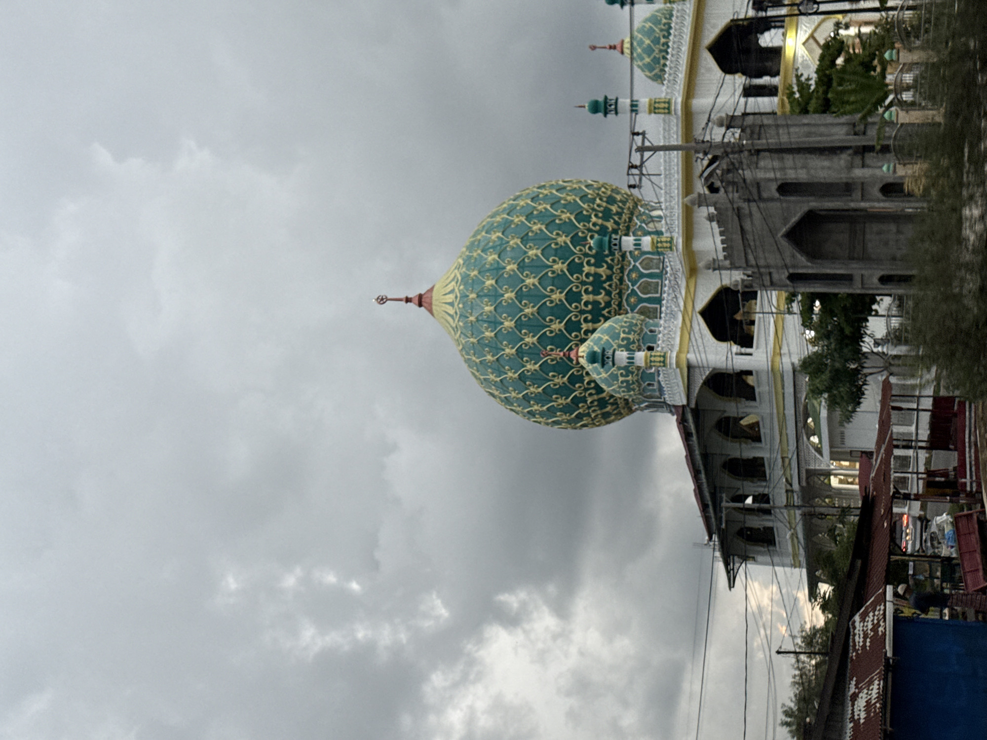 The image depicts the Islamic Center of Lhokseumawe in Indonesia. The focal point is a large, green and gold dome. The building beneath is multi-storied and white. In the foreground are rooftops and wires. The sky is overcast and gray. The image is rotated 90 degrees clockwise.