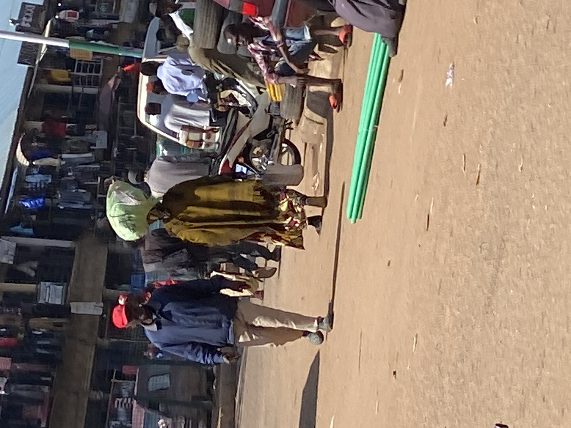 This image captures a street scene in Bukuru, Nigeria, likely a market area based on the surrounding stalls and diverse activities. In the foreground, a man wearing a blue jacket, light-colored trousers, and a red cap walks towards the left of the frame. To his right, another individual, partially obscured, carries a large, light green bundle on their head, wrapped in plastic sheeting. This person is wearing a yellow-brown patterned garment.
The background reveals a bustling environment with structures that appear to be shops or market stalls. Clothes and various items are displayed outside these establishments. Several other individuals are visible in the middle ground and background, engaged in walking or standing. A white vehicle is parked to the right of the frame, and a motorcycle is parked further back. Tires are stacked near a seated man in a red and black checkered shirt, blue jeans, and orange sandals. In the extreme foreground, a cluster of bright green pipes lies on the ground. The overall impression is one of a busy urban market on a bright, sunny day.