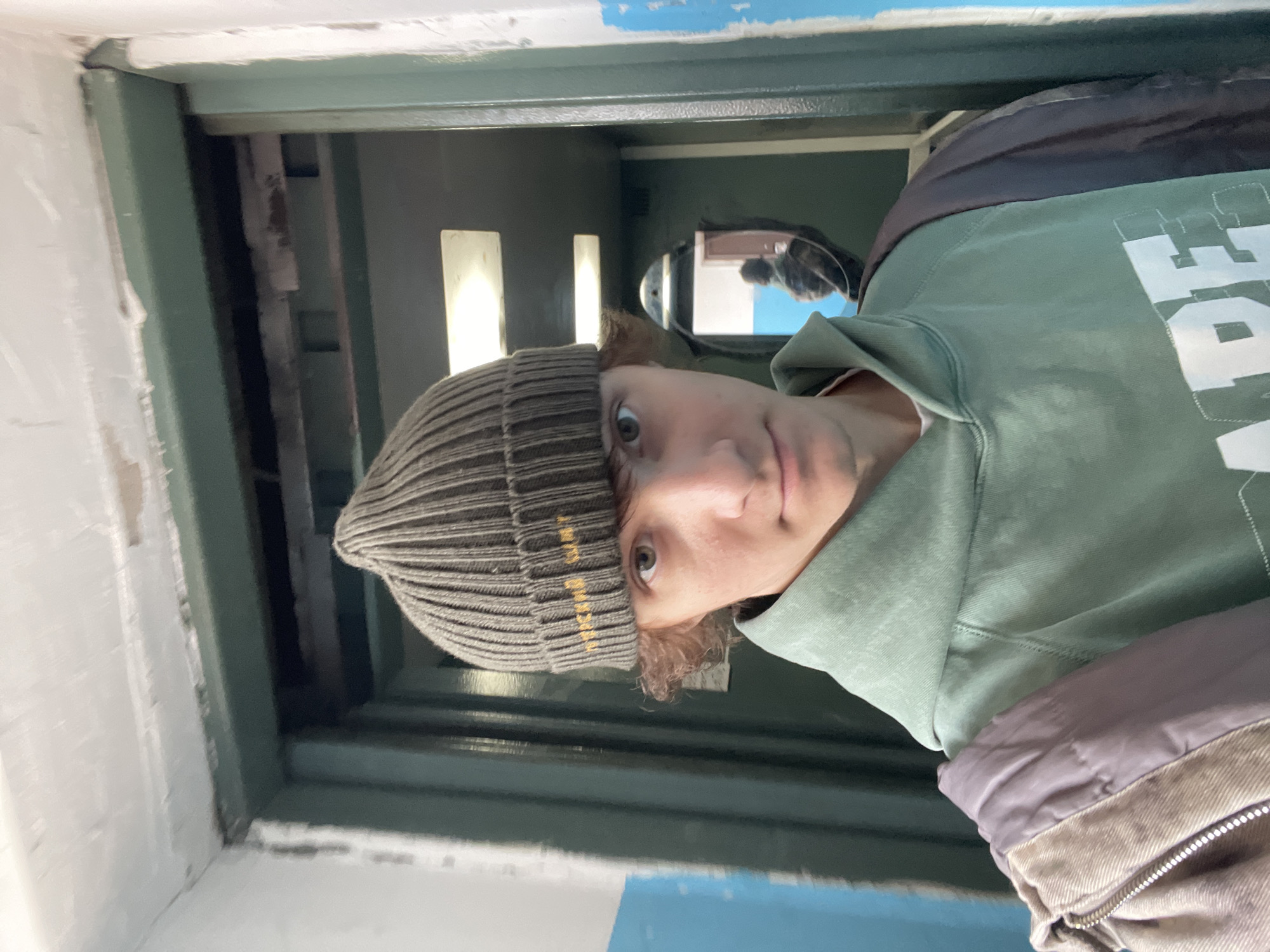 A close-up, slightly angled photo depicts a person indoors, wearing a gray ribbed beanie with yellow lettering, a light green hooded sweatshirt, and a jacket. The person's face is visible, with fair skin, light brown hair, and greenish-brown eyes. The background appears to be an interior space with gray-green walls and white trim. There's a framed opening with bars in the background. The location is possibly in the city of Krasnoyarsk, Russia.