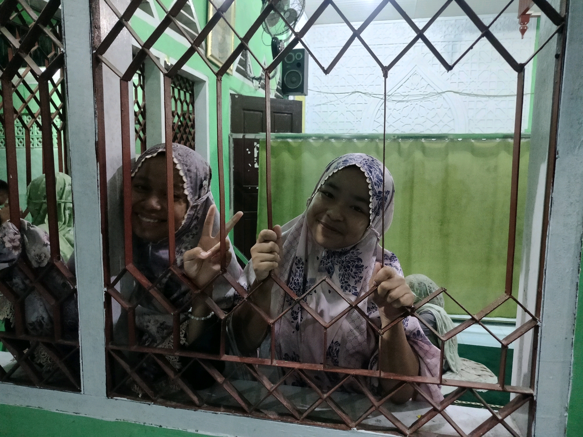 Two young females, wearing headscarves, are visible through a metal grate. The female on the right is smiling and holding the grate with both hands, while making a peace sign with her left hand. The female on the left is also smiling and making a peace sign with her left hand. Behind them, a green curtain is visible, and in the background, a white textured wall with arched patterns can be seen. The scene appears to be indoors, possibly in a building with decorative elements, with the grate suggesting a window or a gate. The overall impression is of a friendly and youthful interaction, possibly in a public space or community building. The presence of a speaker and a fan on the wall suggests the location is a functional space within the city of Purwodadi, Indonesia.