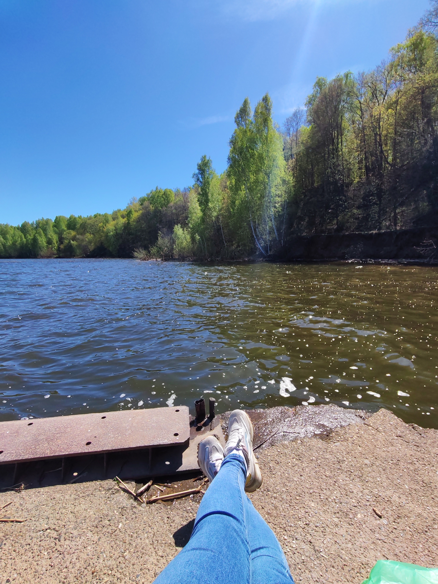 A photograph taken in Икское Устье, Russia, shows a person sitting with their legs extended towards the camera, wearing blue jeans and white sneakers. They are positioned on a concrete platform or dock near a body of water. To the left of the person's legs is a rusty metal structure. The water is dark and reflects sunlight, with visible foam or bubbles near the edge of the platform. In the background, there is a tree-lined shore with lush green foliage under a clear blue sky. The sun is shining brightly.
