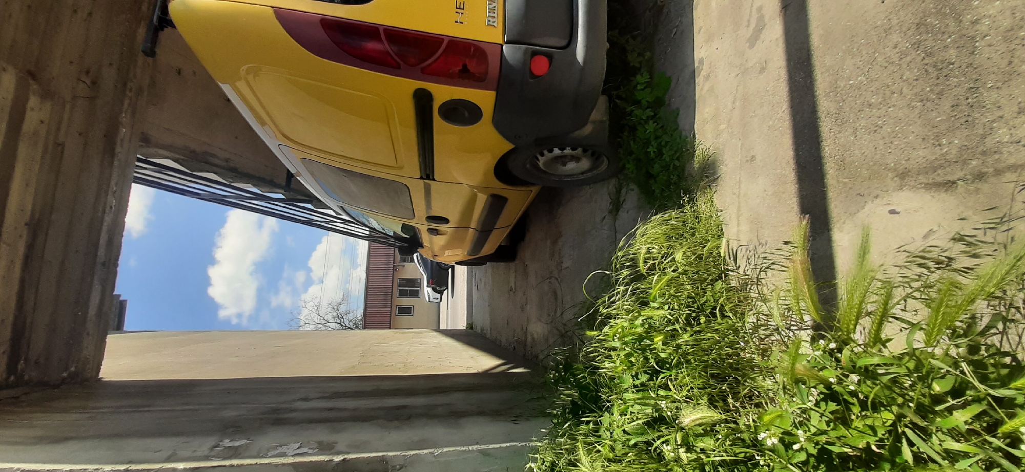 The image depicts a narrow alleyway with a yellow Renault Kangoo van parked on the right side. The van is positioned parallel to a concrete wall on the left, with overgrown green grass and weeds in the foreground, bordering the wall and the van's rear wheel. The alley is covered by an overhead structure with a concrete ceiling and a railing on the right, partially obscuring a bright blue sky with scattered white clouds. In the background, through the opening of the alley, a building with a red roof and two windows is visible, with a white car and a dark-colored car parked on a street in front of it. The scene appears to be an outdoor urban or semi-urban environment. No people are present in the image. The location is identified as Yessentukskaya, Russia.