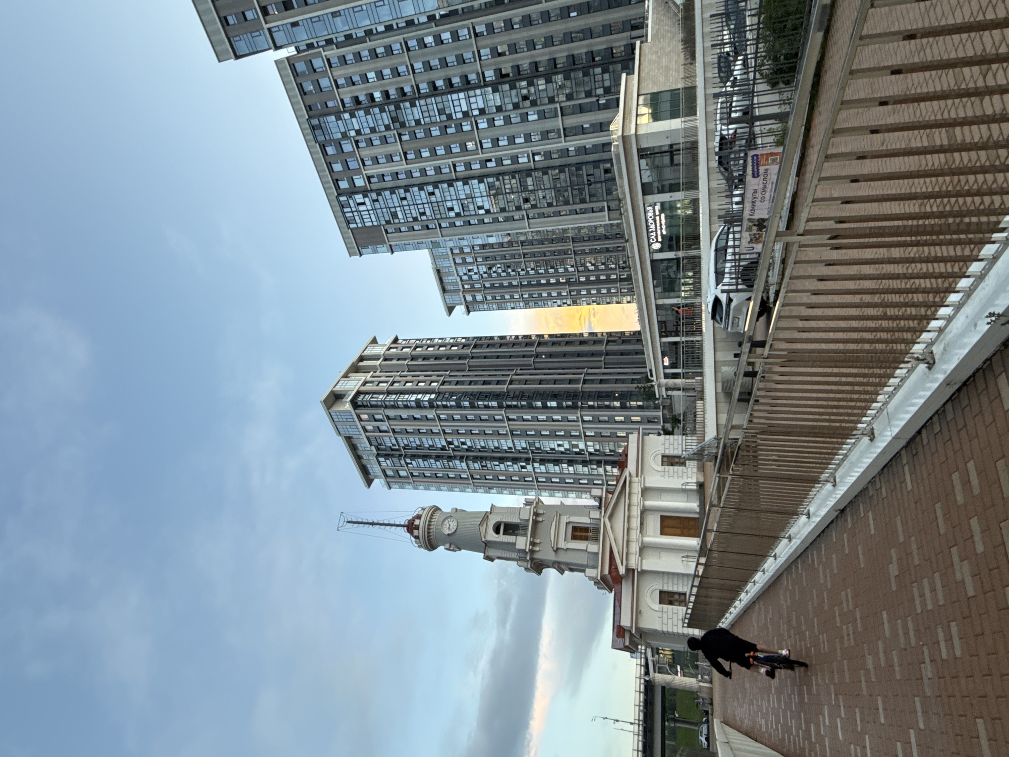 In this eye-level outdoor shot taken in Moscow, Russia, a person is cycling on a brick pathway. The path is bordered by a metal railing, and a modern building complex with numerous windows dominates the background. To the left of the modern buildings, a historical tower with a clock face stands tall. The sky is partly cloudy with hues of blue and hints of orange from the setting sun.
