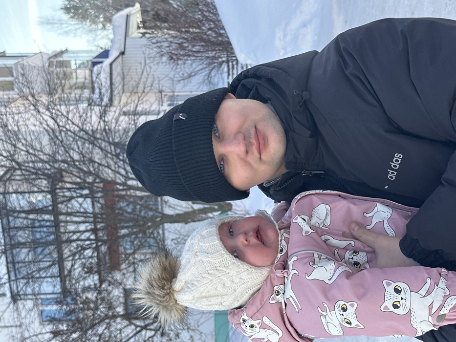 A winter outdoor scene in the city of Chaikovsky, Russia, depicts an adult male holding an infant. The ground is uniformly covered in white snow. The male, positioned on the right, wears a black ribbed knit beanie with a small dark logo and a black hooded puffer jacket featuring the "ad das" logo in white text on the chest. He has blue eyes and a slight smile, looking directly at the camera. He supports an infant, positioned on the left, who is dressed in a cream-colored cable-knit beanie with a brown and white faux fur pom-pom and a pink insulated snowsuit patterned with various white cats. The infant has light-colored eyes and a slightly open mouth, appearing distressed, while also looking towards the camera. In the background, bare deciduous trees are visible in front of residential structures. A multi-story building on the left features large windows reflecting blue light, while a structure to its right has white siding and a snow-covered sloped roof. The scene is brightly illuminated by natural sunlight.