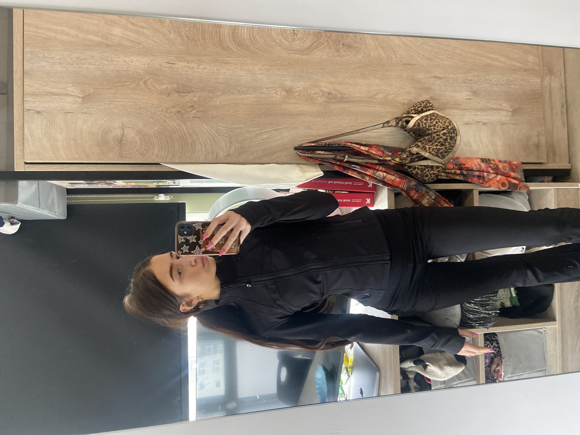 A woman is taking a selfie in a mirror. She is wearing a black top and leggings. Behind her is a cabinet with wood-like doors. There is a leopard print bag and some red and orange patterned fabric on top of the cabinet. The mirror is reflecting a window with buildings outside, suggesting an urban environment. However, without additional information, the exact location in London, United Kingdom, cannot be determined.