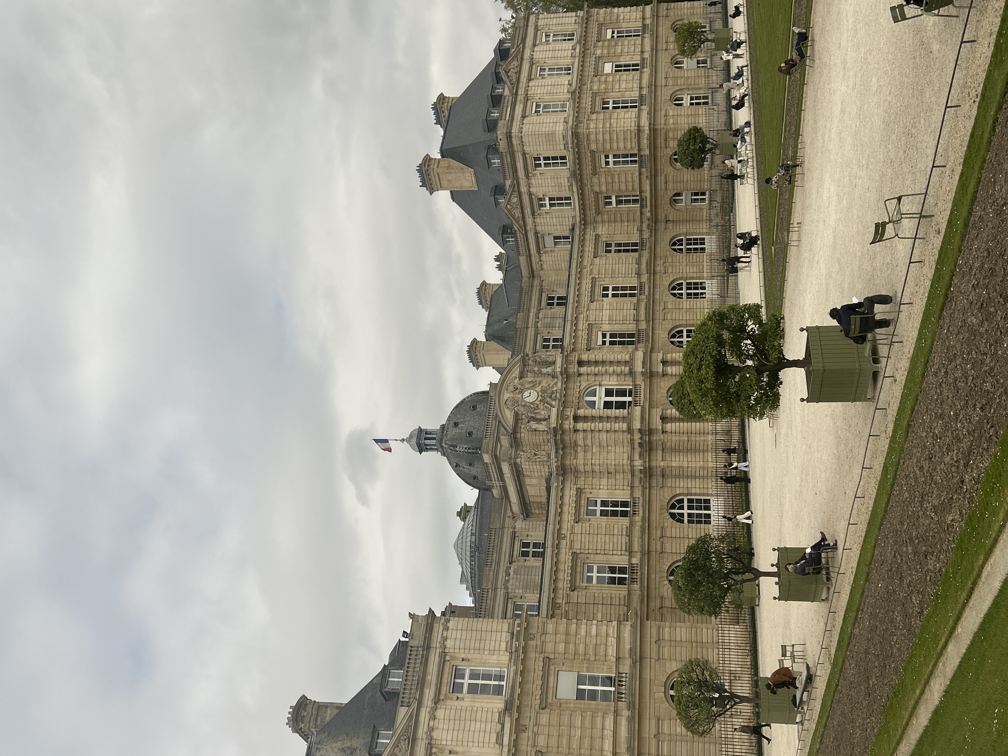 The image depicts the Palais du Luxembourg in Paris, France, under an overcast sky. The palace is a large, multi-story building constructed of light-colored stone with numerous arched windows. A prominent dome with a clock and a French flag atop it rises from the central section of the building. In front of the palace is a gravel path with rows of green metal chairs. People are sitting and relaxing in the chairs. Small trees are planted in green containers along the path. A dark wall or barrier separates the path from a green lawn. The photograph has been rotated counterclockwise.