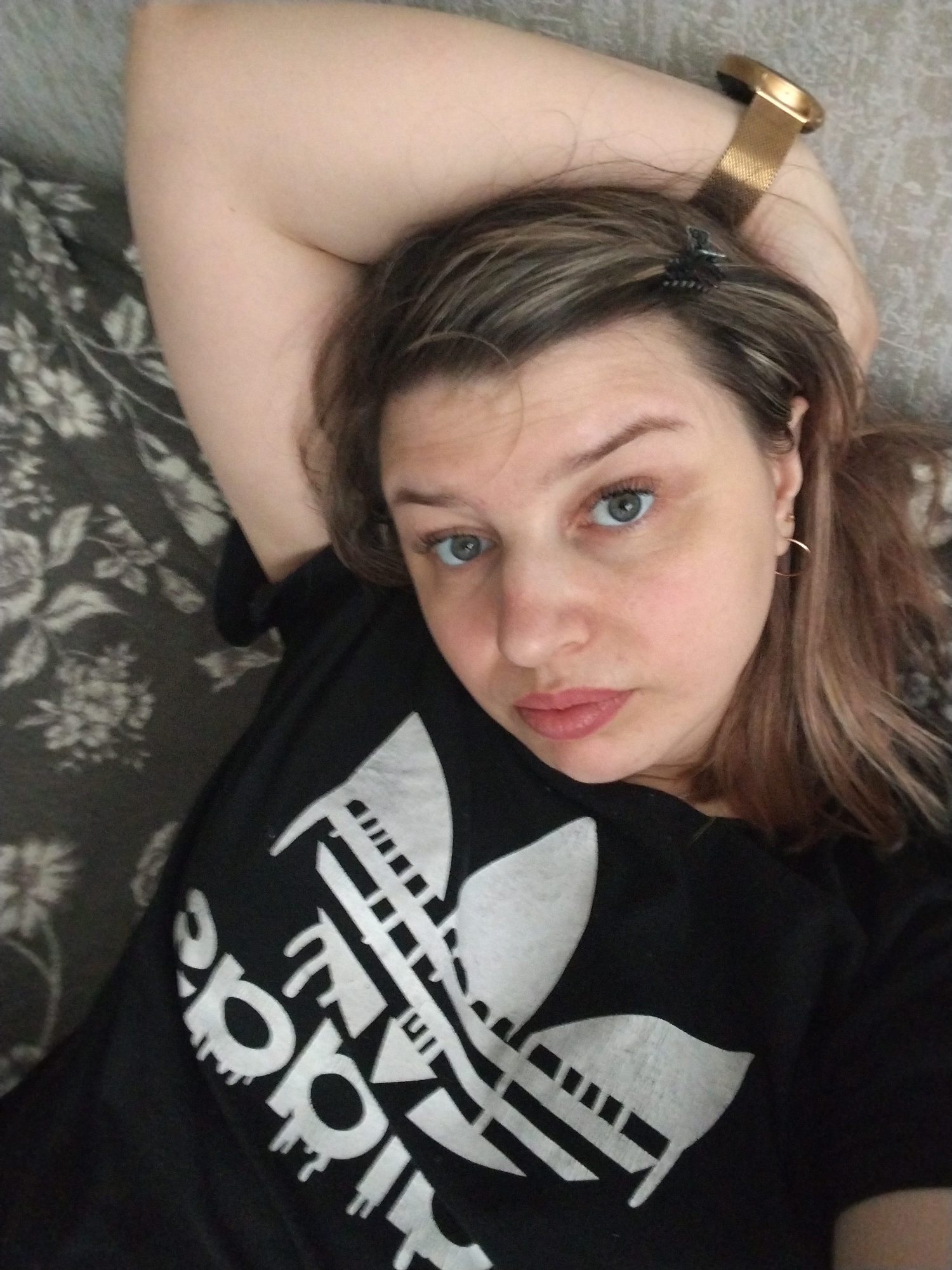 A woman is lying down, likely on a bed or couch, with her left arm raised above her head. She is wearing a black T-shirt with a large white Adidas logo. She has light brown hair with blonde highlights and blue eyes. She is wearing a gold watch on her wrist. A small hair clip is visible in her hair. She is wearing small gold hoop earrings. The background appears to be a floral patterned fabric. There is no information to indicate that the image was taken in Rusanovo, Russia.