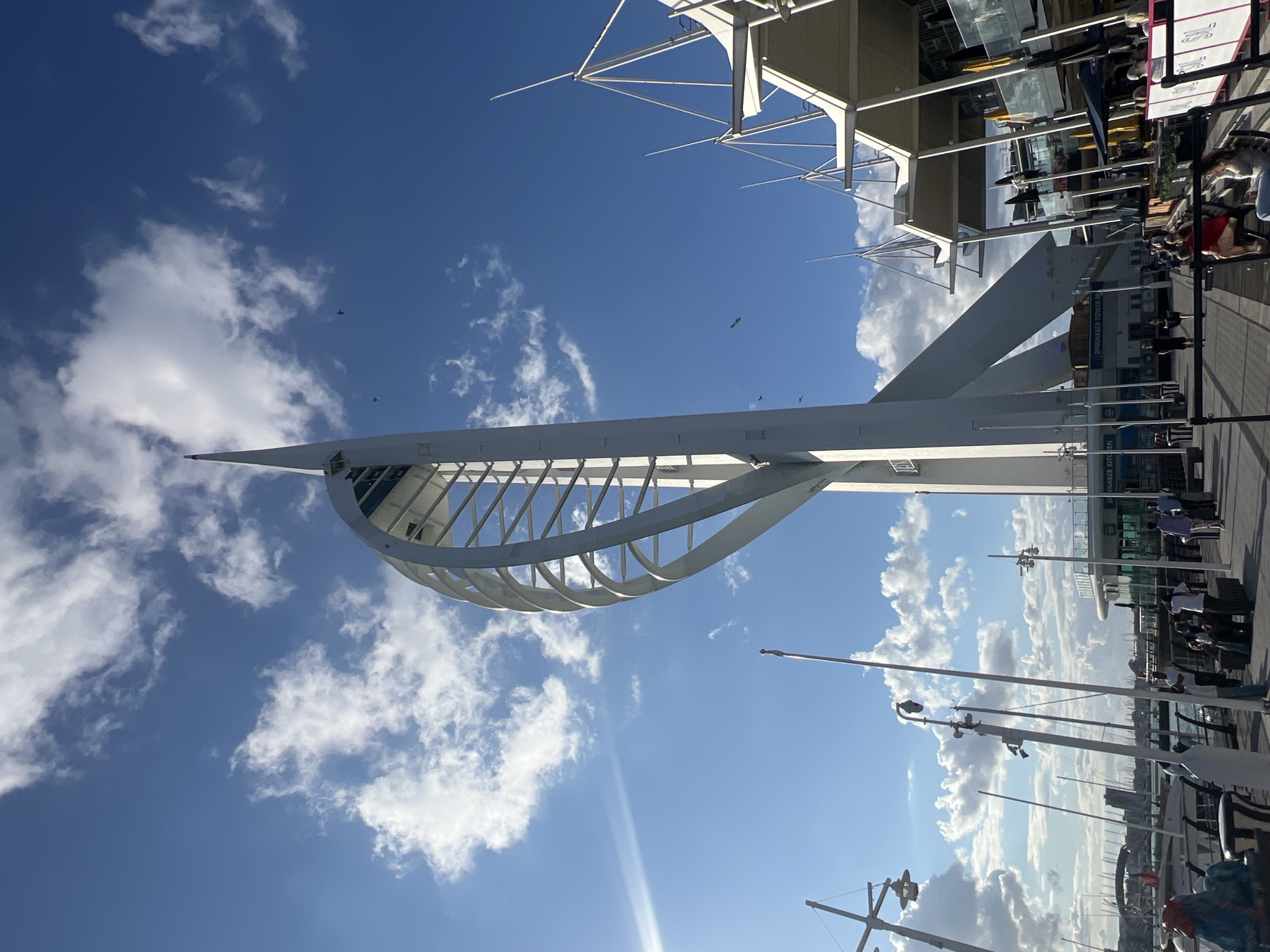 This image captures the Spinnaker Tower, a prominent landmark in Portsmouth, United Kingdom, on a bright, sunny day with scattered clouds. The tower, a tall, white, modern structure with a distinctive sail-like design, dominates the center of the frame. Its sleek, curving white supports spiral upwards, culminating in a pointed spire.
To the right of the tower, a contemporary building with a white, angular roofline and multiple levels of windows is visible, appearing to be part of the surrounding waterfront development. Below this building, outdoor seating areas with tables and umbrellas suggest a bustling commercial or dining area.
The foreground features a paved walkway, a common area for pedestrians. Several people are walking and standing on this walkway, engaged in various activities: some are in small groups, others are walking individually, and some appear to be seated at the outdoor tables. There are also numerous flagpoles and streetlights lining the walkway.
In the background, beyond the walkway and buildings, a body of water is visible, filled with numerous sailboats docked in a marina, indicating the presence of a harbor. The sky is a vibrant blue, with fluffy white clouds creating a dynamic contrast. Two birds are captured in flight against the blue sky, adding a sense of scale and life to the scene. The overall impression is one of a lively, modern urban waterfront setting.