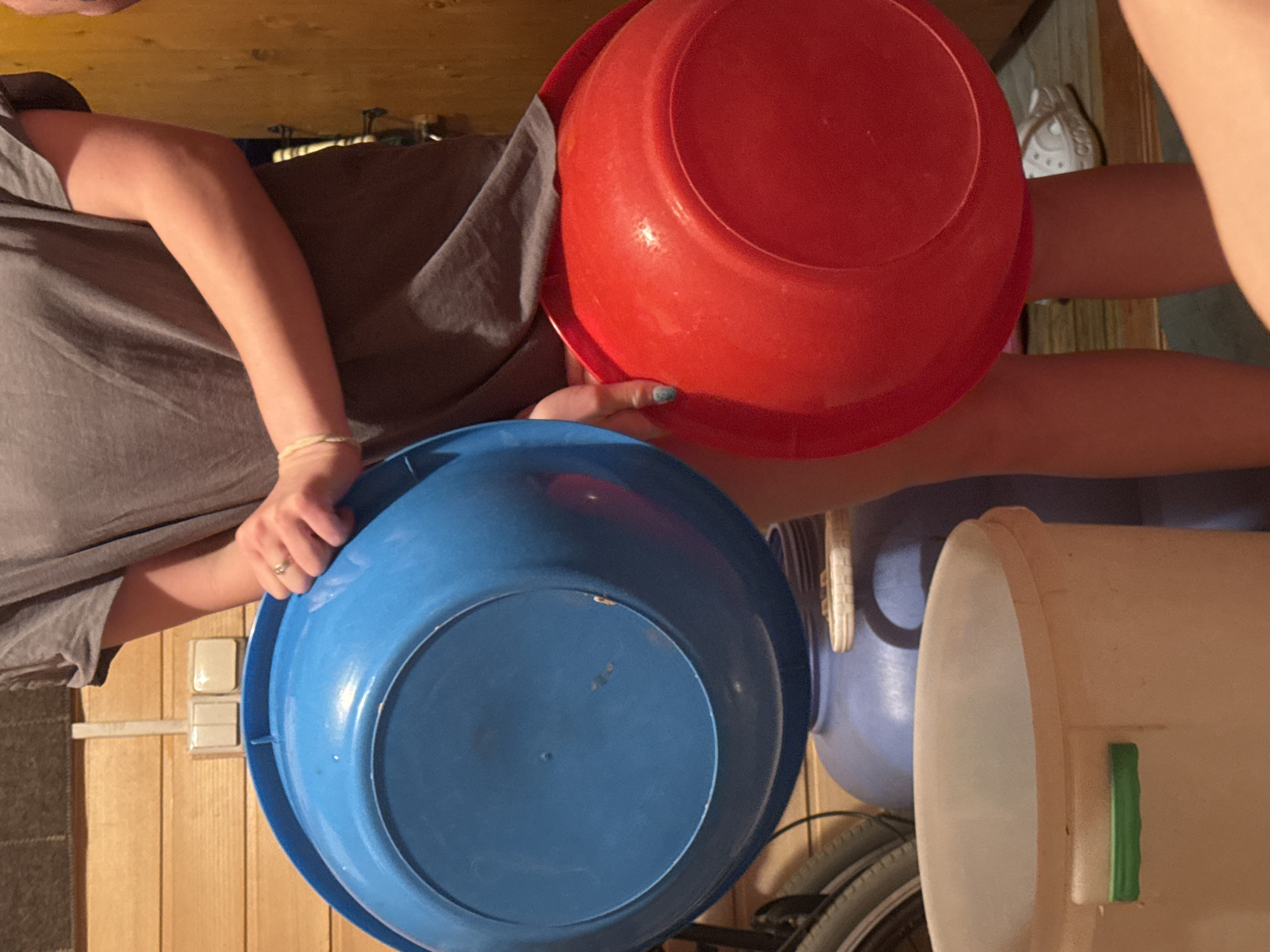 The image shows two individuals holding a blue plastic bowl and a red plastic bowl, respectively. Another blue plastic bucket and a white plastic bucket are visible in the background. The setting appears to be indoors, possibly a storage or utility room, given the wooden walls and the presence of a bicycle wheel and other household items. Details to support a precise location in Mayak, Russia, are absent.