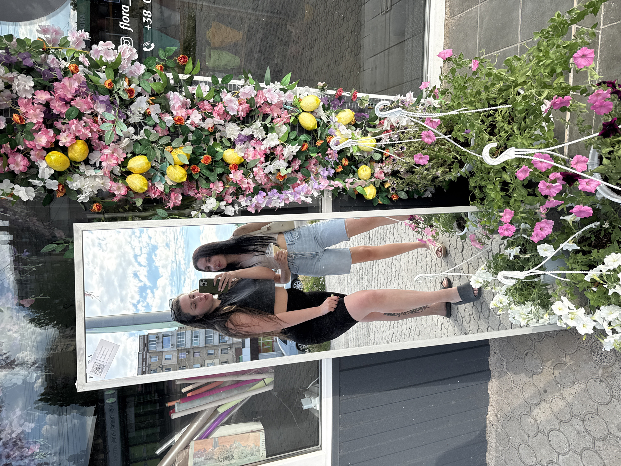 A full-body mirror reflects two women standing outside a flower shop. The woman on the left, with long dark hair and sunglasses perched on her head, wears a dark crop top and black shorts. She is holding a smartphone and posing for a selfie. The woman on the right, also with long dark hair, wears a light-colored top and denim shorts, and holds a drink with a straw. She is looking at the camera. The flower shop is decorated with an abundance of artificial flowers and lemons. The background shows a building and a car. The ground is paved with concrete tiles.