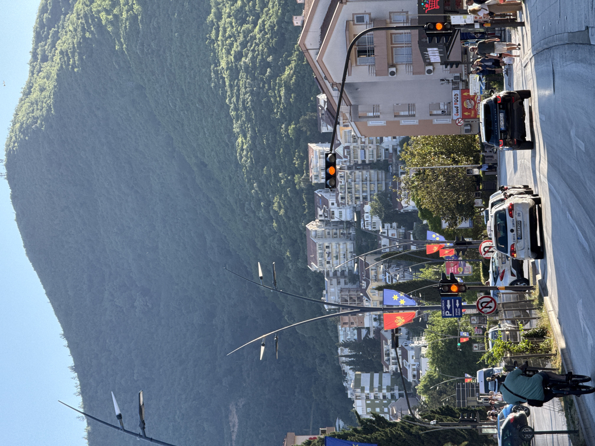 The image shows a street in Budva, Montenegro, with a large, green mountain rising on the left. The right side of the image features a road with cars, traffic lights, and pedestrians. Buildings and houses are visible along the street, nestled into the hillside. The road is lined with flags and street signs. A black SUV is approaching the traffic light. A person is riding a bicycle along the side of the road in the direction the traffic flow.