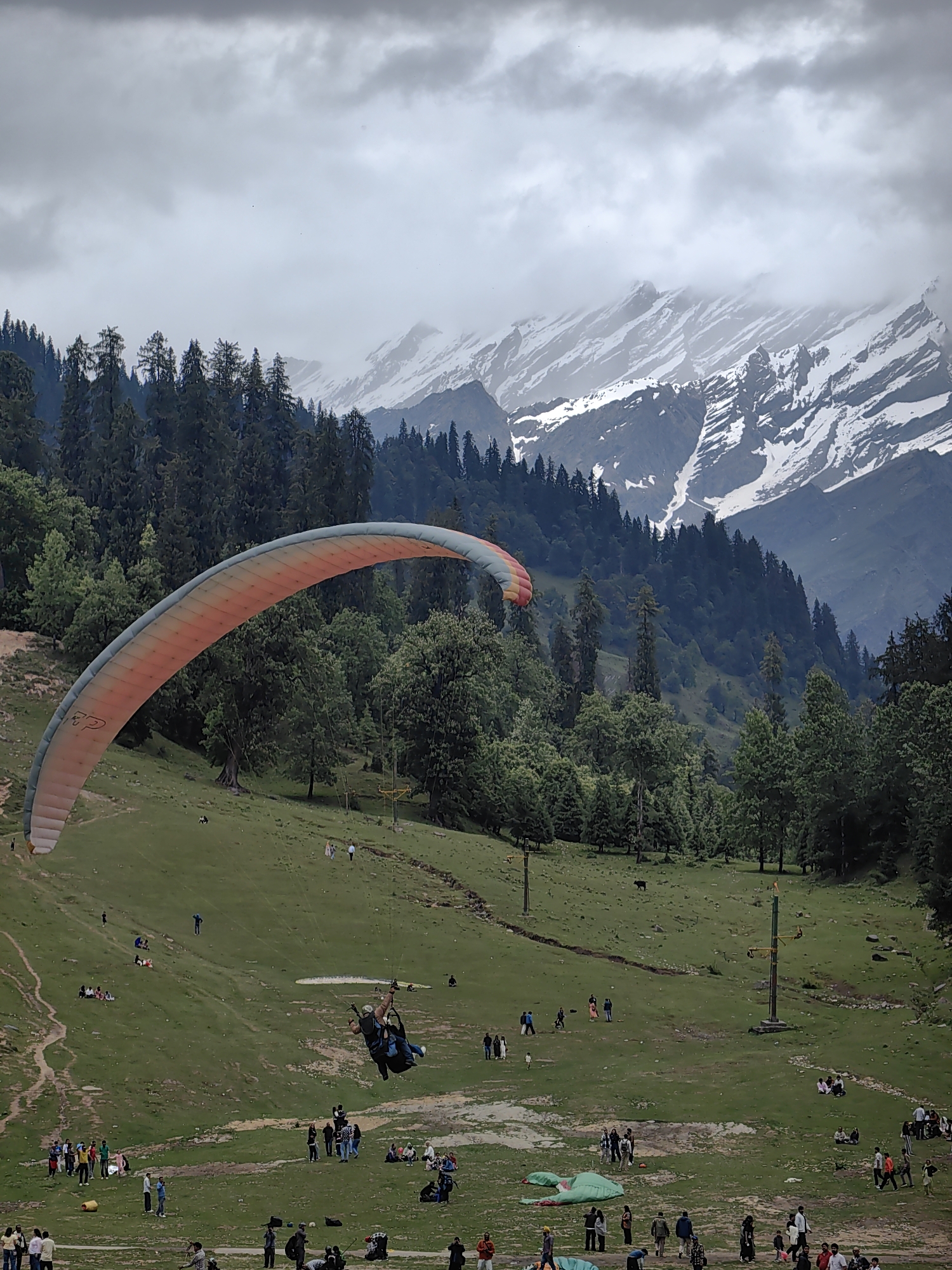 This is a vertical, wide-angle photograph of paragliding in Solang Valley, India. In the foreground, a paraglider with a colorful orange and white canopy is in mid-air, with two figures suspended beneath in a harness. Below the paraglider, a grassy slope is dotted with numerous people, some sitting and some standing. Several tents are visible on the grass, along with a paragliding take-off ramp. In the mid-ground, a dense forest of pine trees covers the hillsides. The background features snow-capped mountains under a cloudy, overcast sky. The overall scene depicts a popular adventure sports destination.