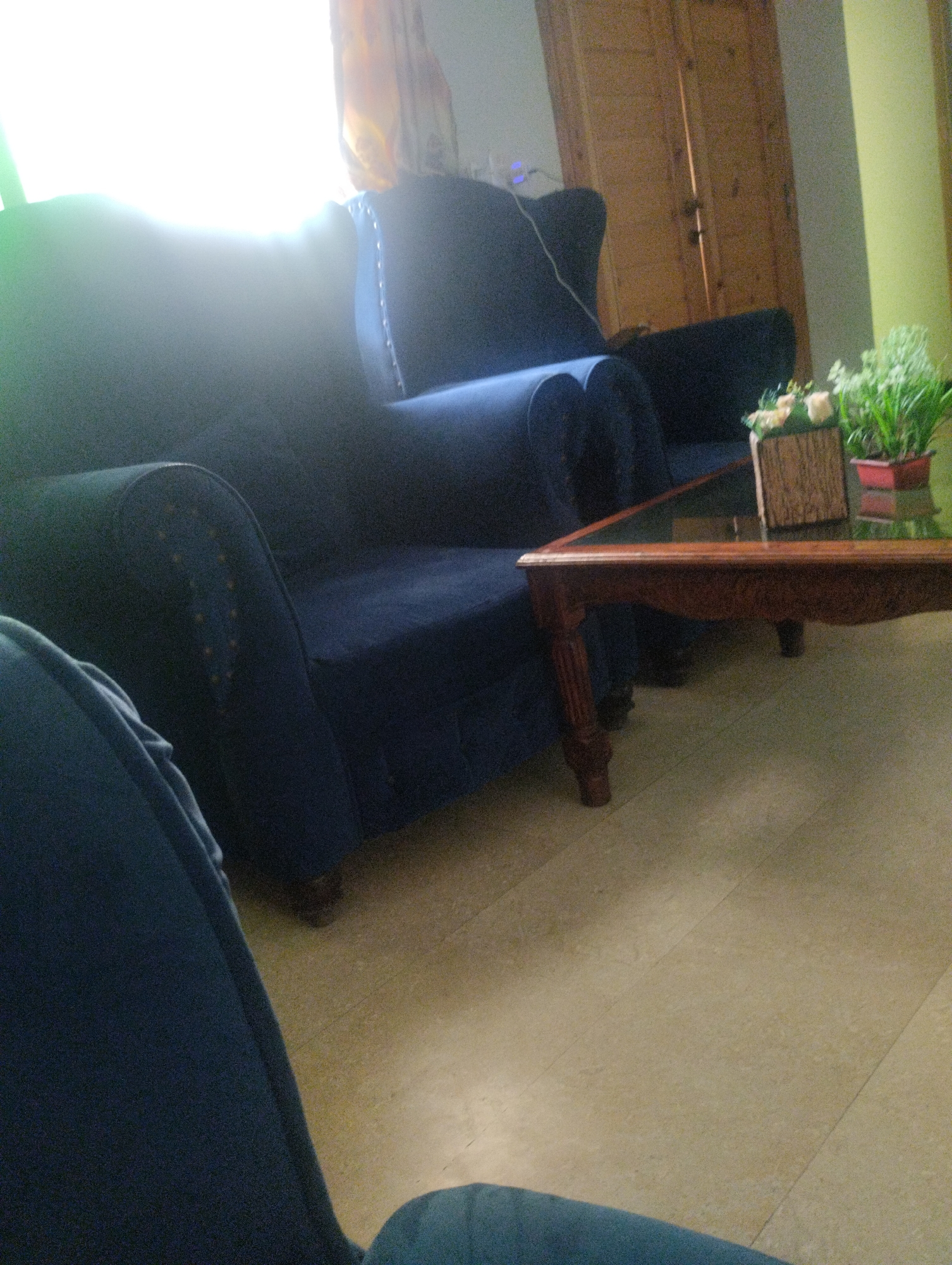 The image shows a dimly lit room with a blue sofa in the foreground. A wooden coffee table with a glass top is placed in front of the sofa. On the coffee table, there is a decorative box and a potted plant. In the background, there are wooden doors and a window with curtains. The floor is tiled. The scene appears to be an interior of a house or a living room.
