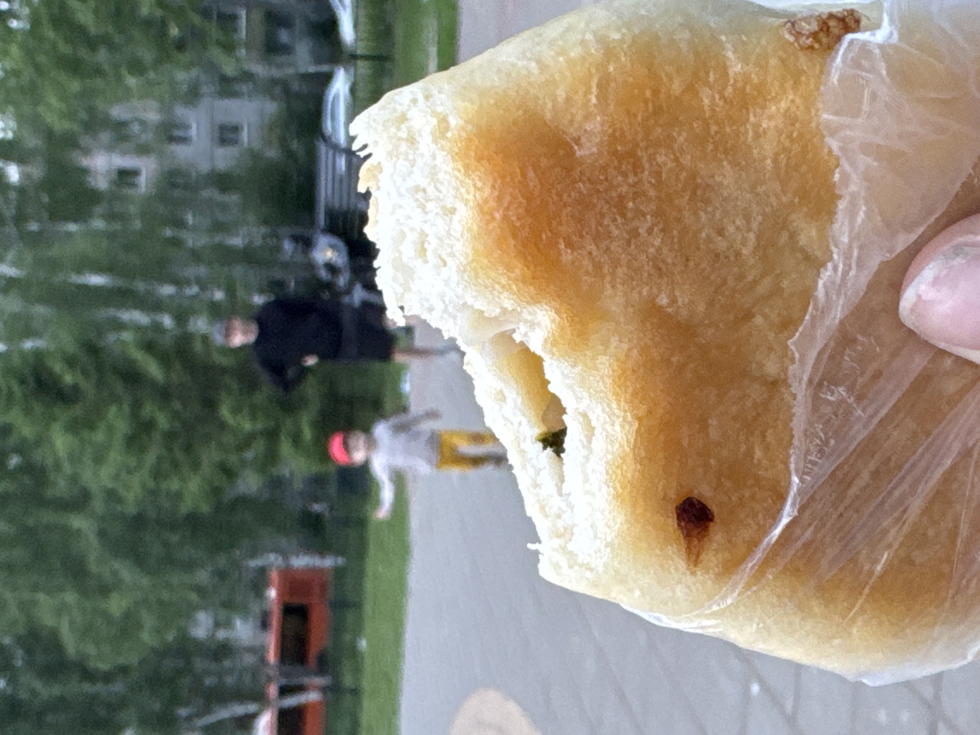 The image depicts an outdoor urban scene in Izhevsk, Russia, with a primary focus on a partially consumed pastry held in the foreground. The pastry, appearing golden-brown with a visible green filling, is enclosed within a transparent plastic bag, with a person's thumb and index finger partially visible at the bottom. In the blurred midground, a young child wearing a red cap, a light-colored long-sleeved top, and yellow pants is captured in motion on a grey paved surface, with both arms slightly extended. Further back, an adult male, dressed in a dark long-sleeved shirt, dark shorts, and a dark cap, stands with hands on hips, observing the scene. The background reveals an abundance of green trees and foliage, partially obscuring residential buildings, including a light-colored, multi-story structure. A red vehicle, likely a bus or trolleybus, is discernible on the far left behind the trees. A dark park bench and additional vehicles are visible in the indistinct distance on the right. The lighting indicates an overcast daytime environment within a park or residential area.