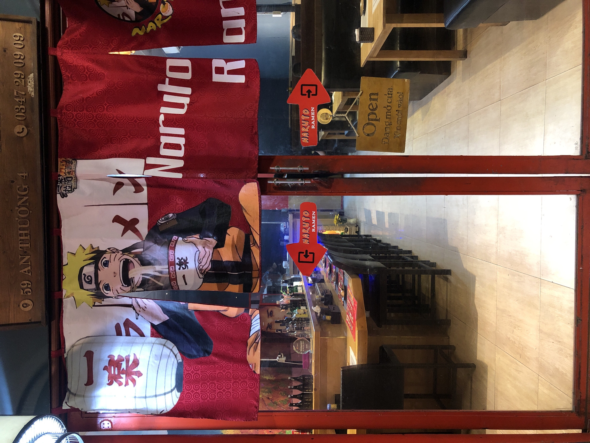 This image shows the entrance to a Naruto-themed ramen restaurant in Da Nang, Vietnam. The entrance is framed by red metal bars and features two doors, one of which is partially open. On the left door, there are red banners with white text that reads "Naruto" and a stylized image of Naruto Uzumaki, the main character from the anime and manga series. A white lantern with red Japanese characters is also visible on this door.
On the right door, a red arrow-shaped sign points downwards and says "NARUTO RAMEN". Next to it, a wooden sign reads "Open" in English and "Đang mở cửa. Xin mời vào!" in Vietnamese, indicating that the restaurant is currently open and customers are welcome.
Through the open doorway, a bar area with shelves stocked with bottles is visible. The interior lighting appears to be a mix of natural light and interior lights, casting reflections on the glass doors. The floor inside appears to be tiled.
The scene suggests a casual dining establishment, likely busy during the day, given the signage. The overall impression is a vibrant and welcoming atmosphere, capitalizing on the popularity of the Naruto franchise to attract customers. The time of day is not explicitly discernible, but the lighting suggests it could be daytime.
