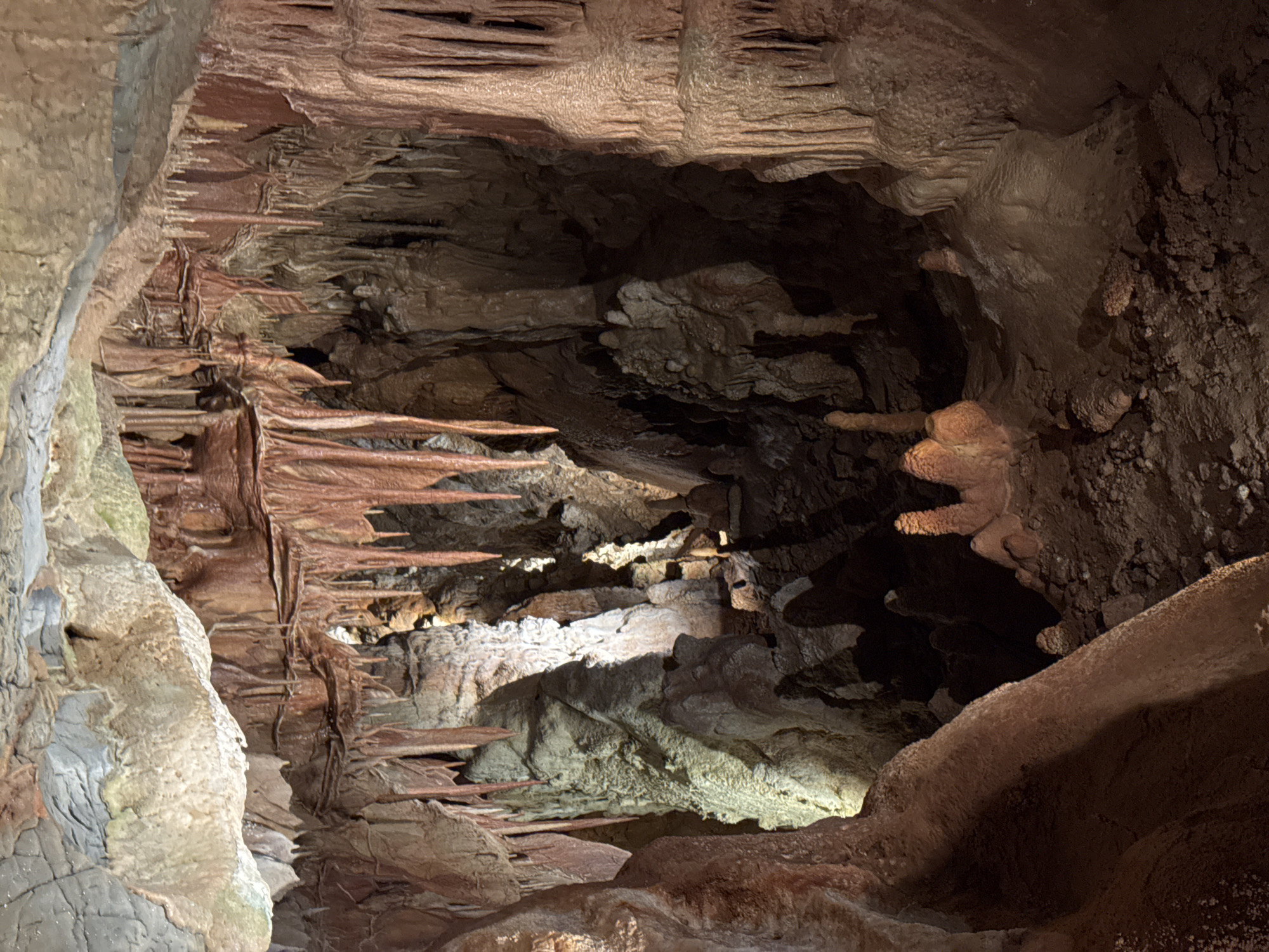 The image shows the interior of a cave system. The walls and ceiling are composed of rough, layered rock formations, with varying textures and colors ranging from light gray to dark brown and reddish hues. There are several speleothems, including stalactites hanging from the ceiling and other formations protruding from the walls. A light source illuminates part of the cave floor and walls, creating strong shadows and highlighting the uneven terrain. The location is Unknown, Unknown. There are no people visible in the image.