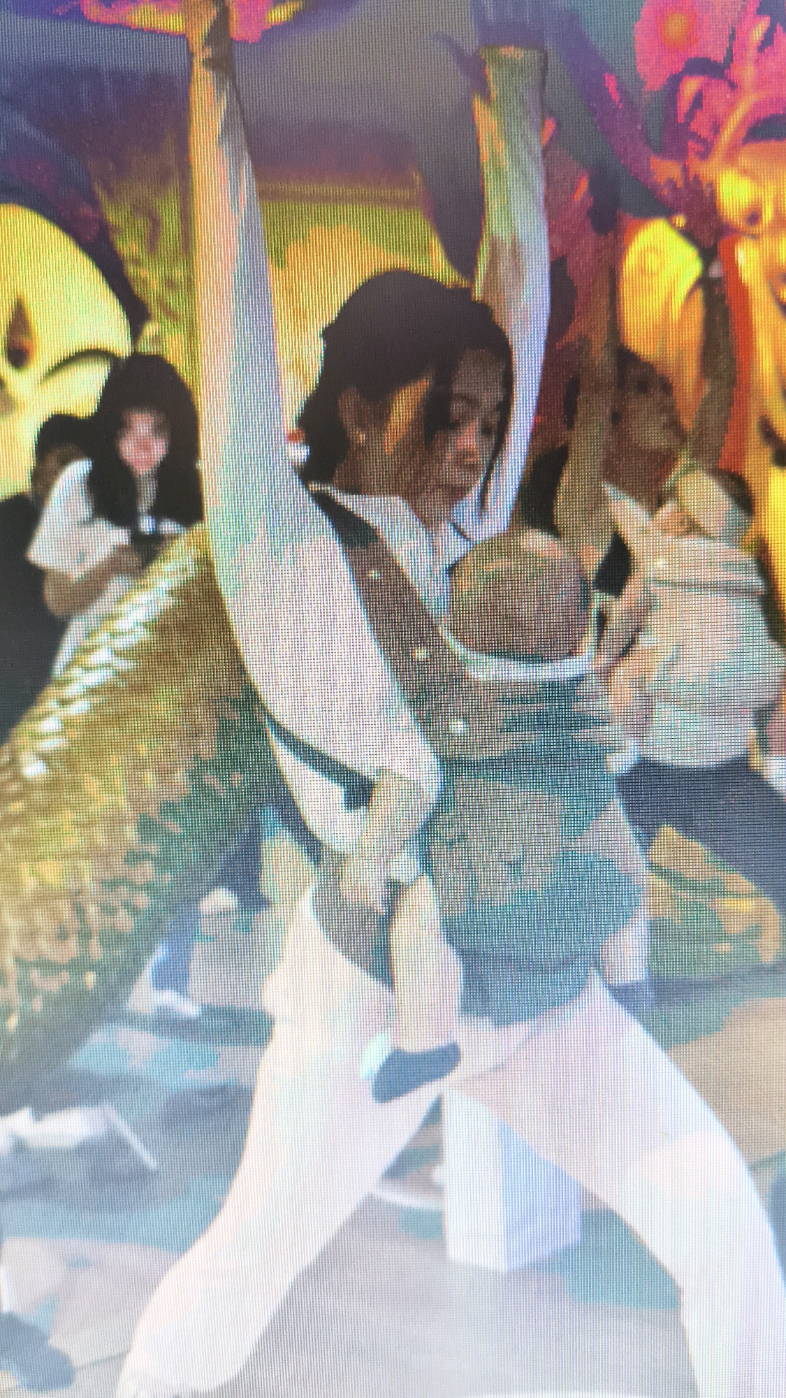 In Setiamekar, Indonesia, a medium shot captures a woman standing amidst a blurred background with colorful patterns, possibly a mural or decoration. She is wearing a white long-sleeved shirt and white pants, with a baby carrier holding an infant strapped to her front. The baby is facing outwards and appears to be wearing a dark-colored outfit. The woman's right arm is raised upwards, while her left arm is bent, with her hand resting on her hip. Her head is tilted downwards. To her left, another person, possibly a female, with dark hair is visible, wearing a white top. Further in the background, the forms of other individuals and elements are indistinct due to the blur. The overall lighting suggests an indoor or dimly lit outdoor setting with vibrant, possibly artificial, lighting contributing to the colorful background.