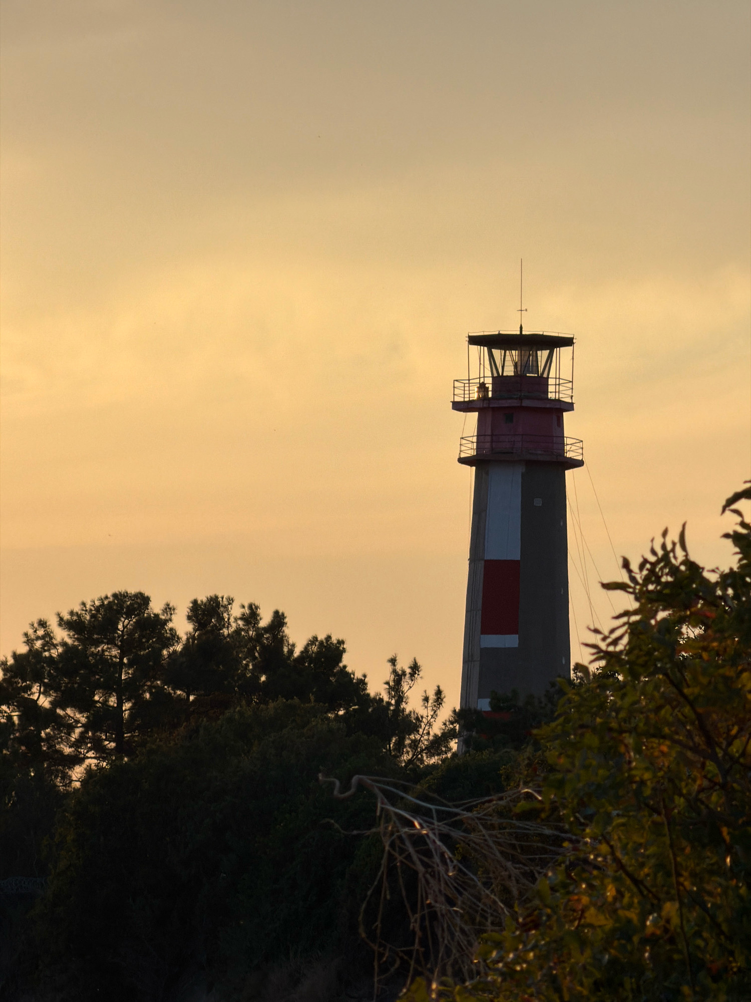 The image shows a lighthouse on a cloudy, orange-tinted sky. The lighthouse is tall and cylindrical, with red and white markings on its upper section. It is surrounded by trees and foliage, suggesting it is located in a natural or park-like setting. The lighting indicates it may be sunset or sunrise. The lighthouse is likely the Gelendzhik lighthouse, a prominent landmark in Gelendzhik, Russia, known for its distinctive design and coastal location. There are no people or specific actions depicted in the image, focusing solely on the structure and its environment.
