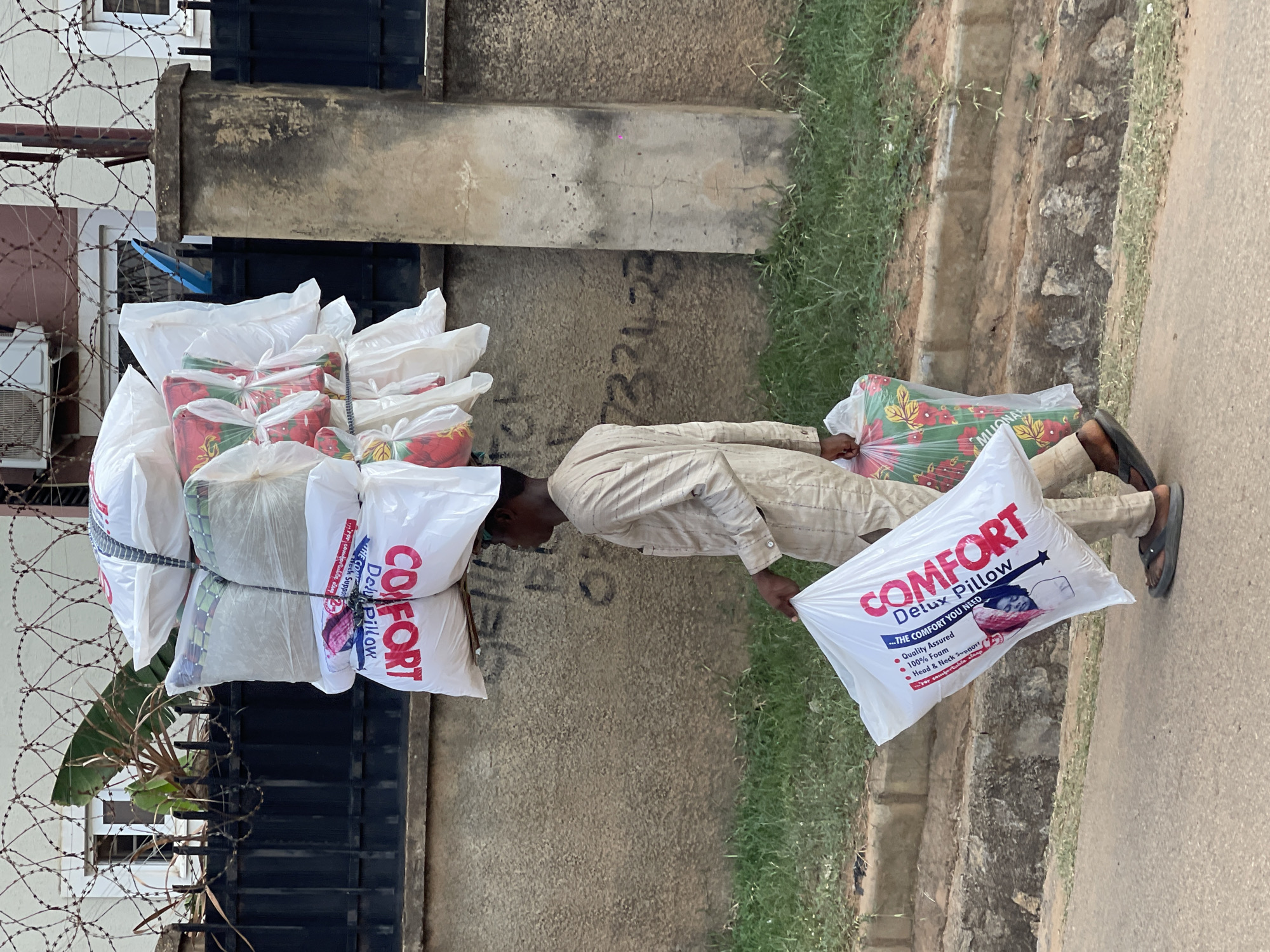 A man wearing a beige shirt and pants is carrying a large stack of "Comfort Deluxe Pillow" brand pillows on his head and one in his hand. He is walking along a sidewalk next to a concrete wall and a patch of grass in a residential area of Abuja, Nigeria. A building with barbed wire and an air conditioner is visible behind him.
