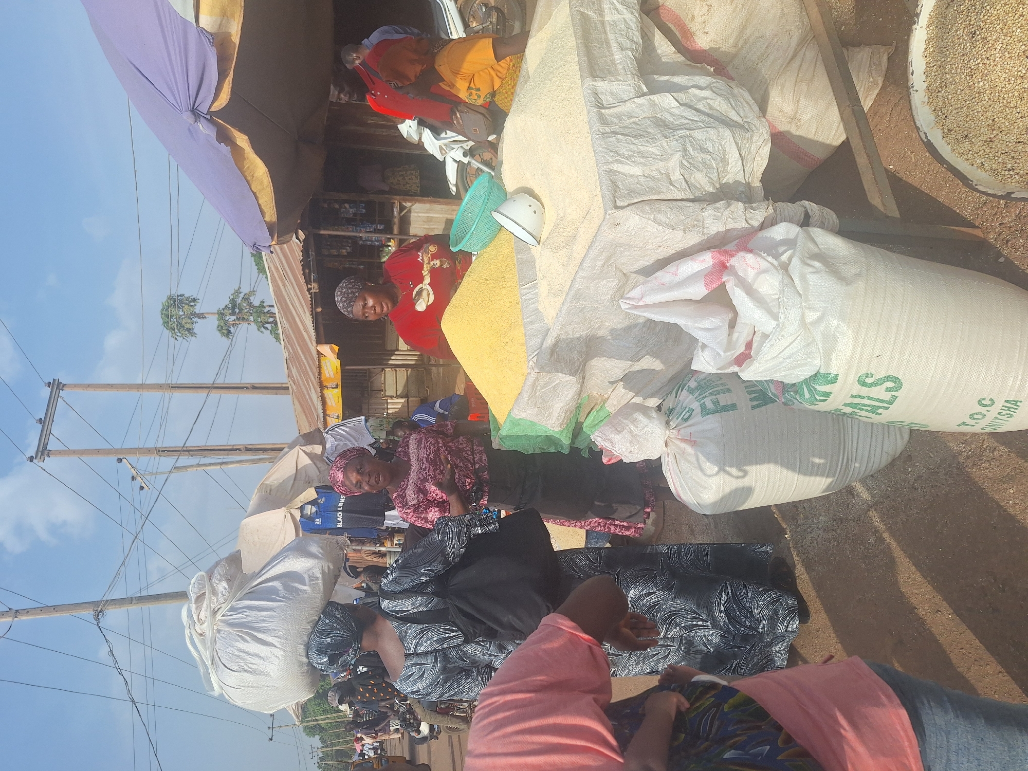 The image captures a bustling marketplace scene in Kakuri Gwari, Nigeria. Several women are visible, interacting with goods displayed in large sacks on the ground. One woman wears a red top and another a pink top. One person is wearing a headscarf with black and white design. There are at least four sacks in the scene. Other objects visible include a blue plastic bowl, a white metal container, a wooden stick, and various bags containing what appear to be grain. The background includes utility poles and wires, a two story building, and other people engaged in market activities. The overall impression is one of a vibrant and crowded outdoor market, characteristic of many urban settings in Nigeria.