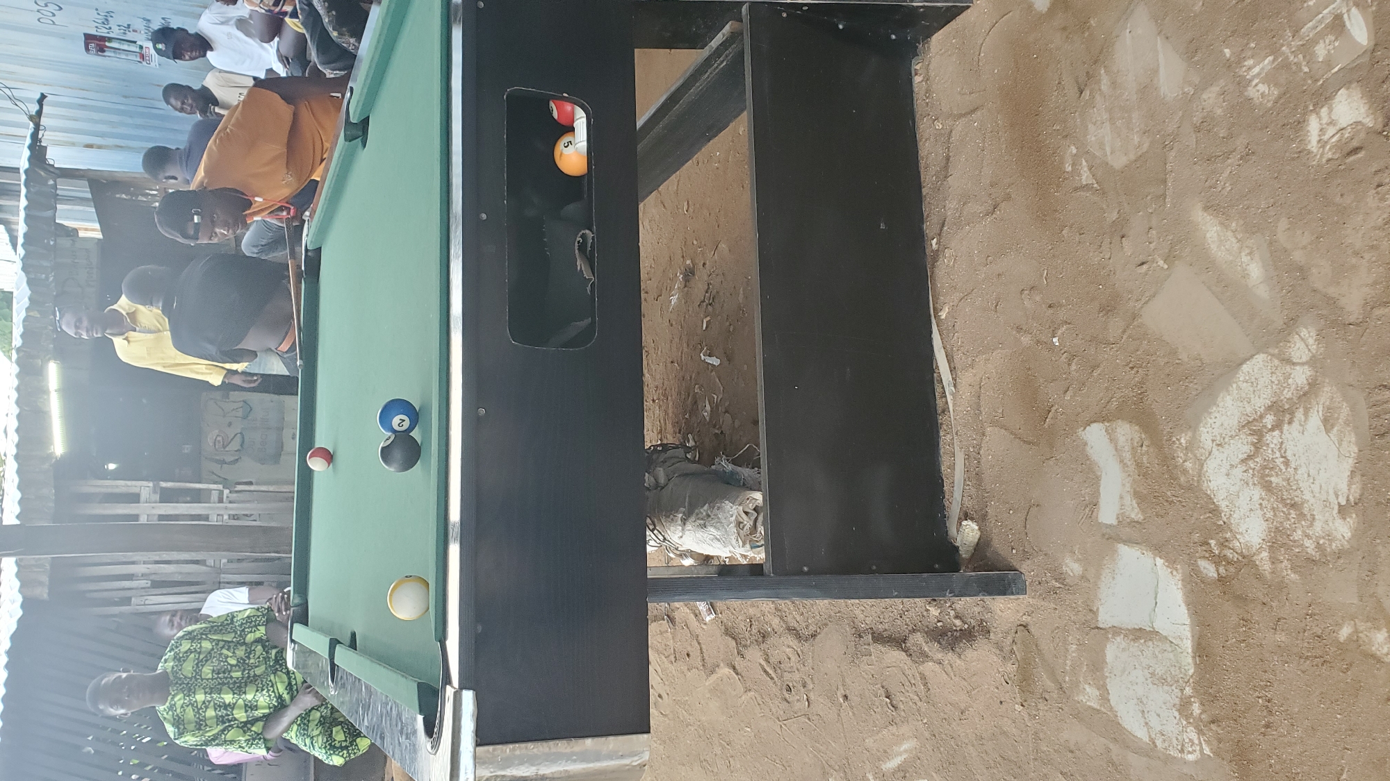 In Jalingo, Nigeria, a group of men are gathered around a green felt pool table. The table is set up outdoors, on sandy ground with scattered debris and some broken white tiles. In the foreground, the pool table has several balls on it, including a white cue ball, a black and gray ball, and a blue ball with the number 2. A red ball is also visible. To the right of the table, a small rectangular opening reveals a pool ball with the number 5 and another red ball. Several men are standing and sitting around the table. One man in a vibrant green and yellow patterned garment is seated on the left. Another man in a yellow shirt stands behind the table. A man in an orange shirt is leaning over the table, appearing to play. Other men are partially visible in the background, seated or standing. The environment suggests an informal gathering place, possibly a bar or outdoor recreational area, with makeshift structures and corrugated metal roofing visible in the background.