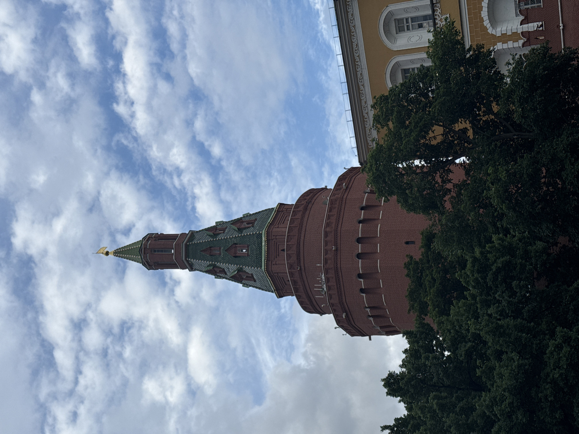 The image shows a view of the Spasskaya Tower of the Moscow Kremlin, located in Moscow, Russia. The tower is a prominent red brick structure with a green, multi-tiered roof topped with a golden spire. The tower's upper section features decorative elements and windows. In front of the tower, a dense canopy of dark green trees obscures the lower part of the structure. To the right of the tower, a section of a yellow building with arched windows and decorative white trim is visible. The sky above is a mix of blue sky and scattered white clouds. The image is taken from a low angle, looking up towards the tower and the sky. There are no people or animals visible in the scene.