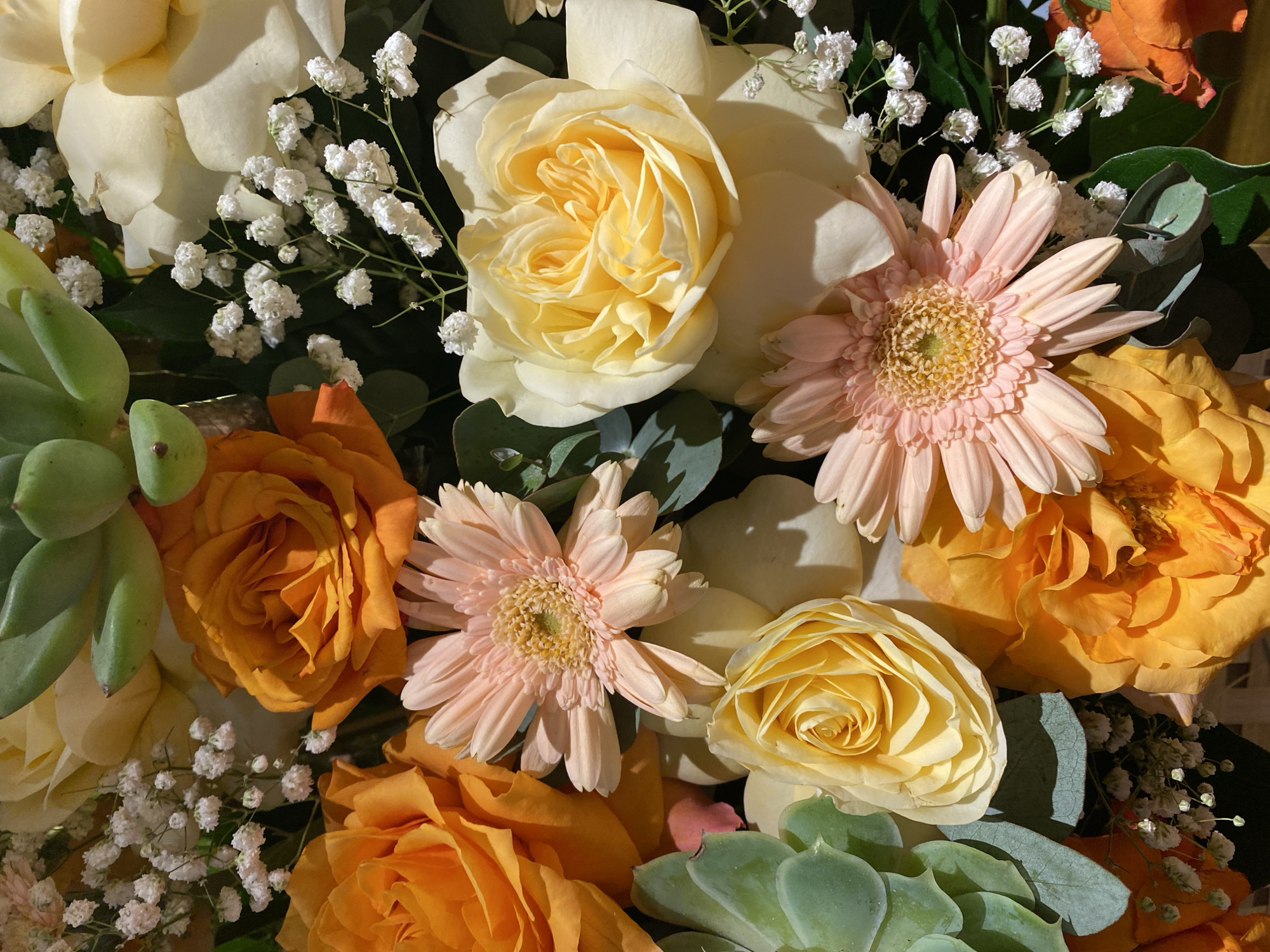 This image depicts a close-up, overhead view of a floral arrangement. The bouquet contains a variety of flowers, including orange and yellow roses, pink gerbera daisies, white baby's breath, and green succulents. The lighting suggests an indoor setting, possibly a home or event space. There are no people or animals visible in the image. The composition is tightly framed, focusing solely on the textures and colors of the flowers. The image does not provide any information that would allow for the identification of a specific location in Đà Nẵng, Vietnam.