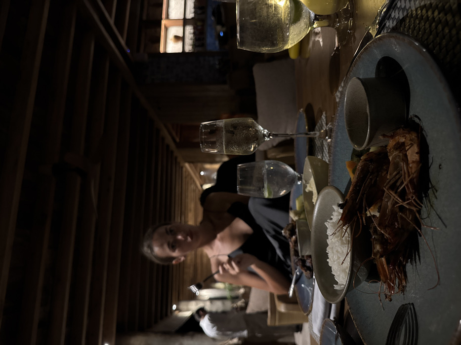 A dimly lit image shows a woman seated at a table laden with food and beverages. Two wine glasses containing sparkling liquid are visible near a plate filled with cooked prawns and a bowl of white rice. Another glass is present, partially obscured, containing a similar beverage. The woman is facing the camera, holding a fork, seemingly about to eat. In the background, there is a blurry figure of a person in a white shirt. The overall setting appears to be an outdoor dining area with a wooden structure.