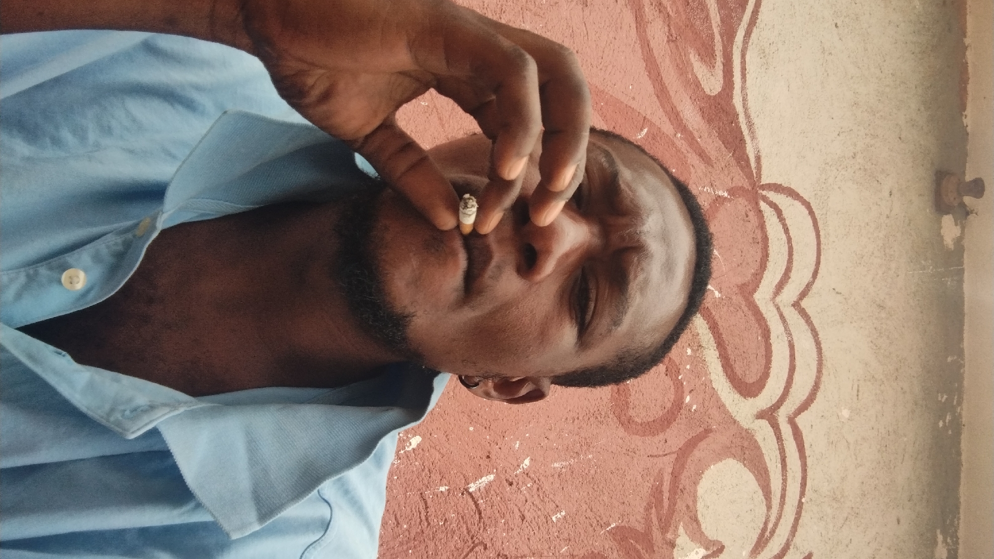 An image, rotated 90 degrees clockwise, features a man of African descent, likely in Jos, Nigeria, in what appears to be a casual setting. The man is seen from the chest up, with his head positioned on the right side of the frame. He has dark skin, a short beard around his chin and jawline, and short dark hair. He is wearing a light blue collared shirt, with the top button unfastened.
The primary activity depicted is the man smoking a cigarette. He holds the lit cigarette between his thumb and index finger, bringing it to his mouth. The cigarette has a visible filter and a small amount of ash at its tip, indicating it has been lit and smoked. His eyes are partially closed, suggesting a relaxed or contemplative state while smoking.
The background consists of a wall with a distinctive decorative pattern. The wall is painted in shades of reddish-brown and off-white or light cream. An ornate, flowing design, possibly abstract or floral, is outlined in reddish-brown against the lighter sections of the wall. There are visible signs of wear and tear on the wall, including chipped paint and faded areas. In the top right corner of the frame (if oriented correctly), a small, dark, cylindrical object, possibly a wall hook or fixture, protrudes from the wall. The lighting suggests it is daytime, but there are no strong shadows to indicate direct sunlight, possibly an indoor or shaded outdoor location. There is no visible text in the image.
