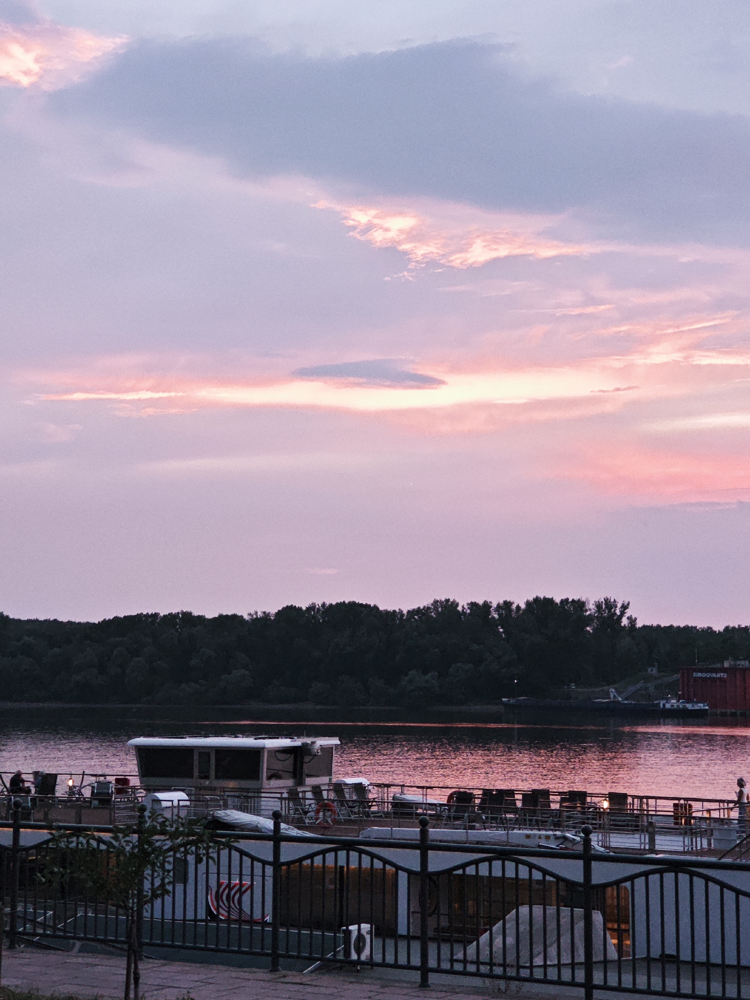 A horizontal, eye-level, medium shot captures a serene river scene at dusk. The sky is a canvas of soft purples and pinks, with streaks of light reflecting off the water. A dark, dense line of trees forms the horizon on the far bank of the river. In the foreground, a white boat is docked, its upper deck adorned with empty chairs. A black metal fence runs along the bottom of the frame, separating the viewer from the boat. A single, small tree stands near the fence. The water is calm, with a shimmering reflection of the sunset. The overall atmosphere is peaceful and tranquil.