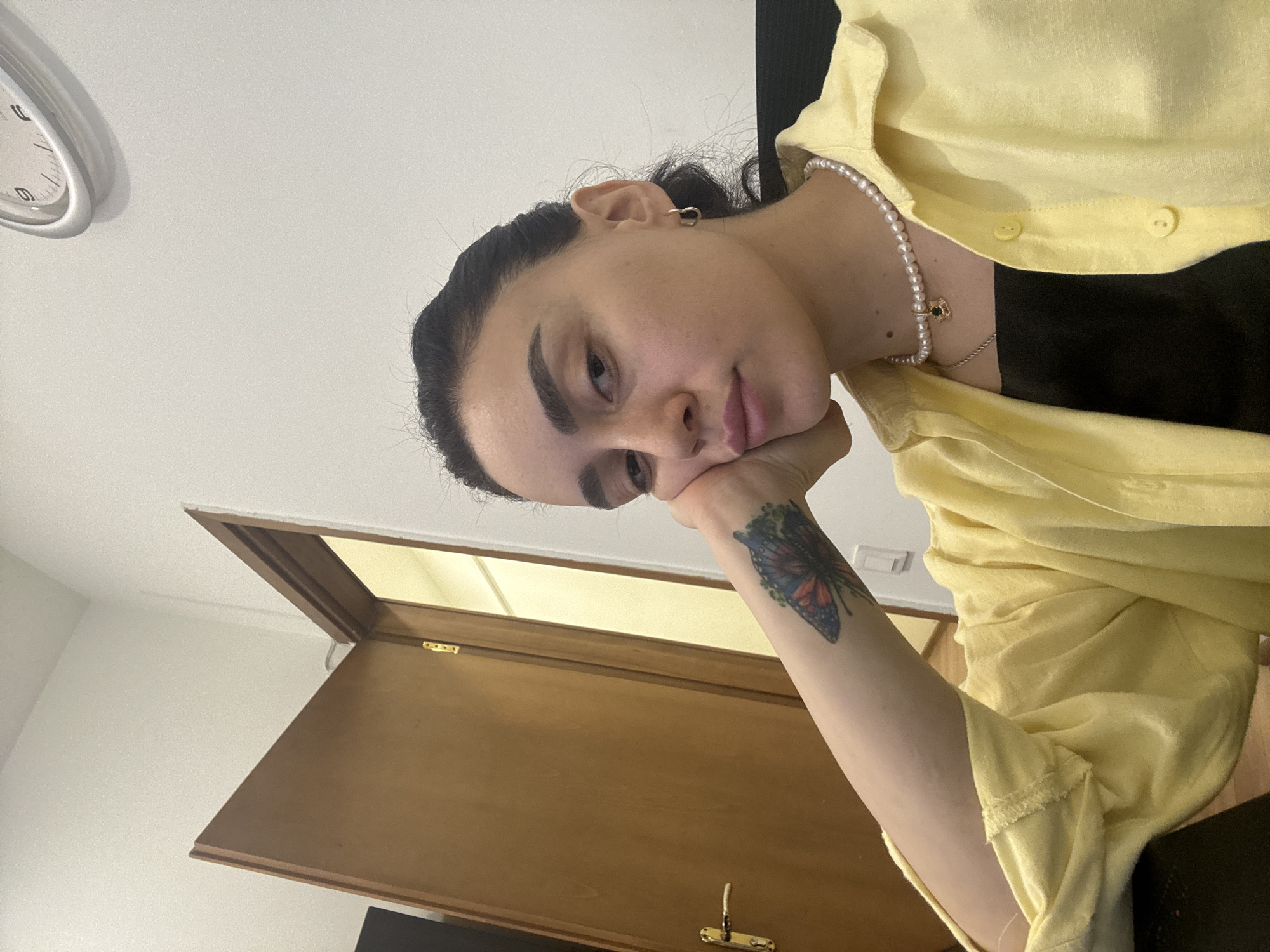 A person with dark hair pulled back and a butterfly tattoo on their right forearm rests their head on their hand. They are wearing a yellow button-down shirt over a black top, and a pearl necklace with a small pendant. In the background, a white wall is visible with a clock on it, and a partially open wooden door. The location is not discernible from the image.