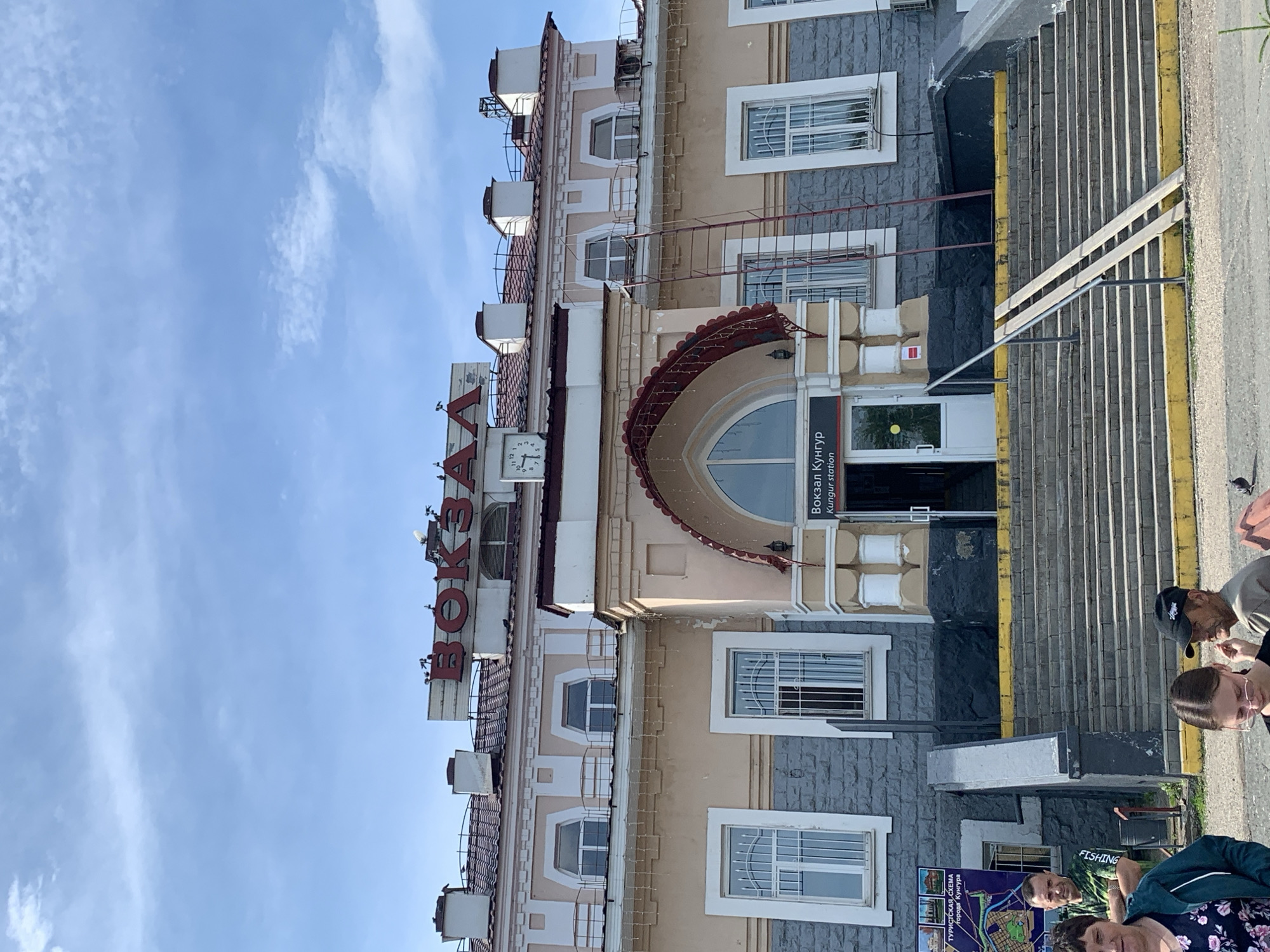 The image depicts the exterior of the Kungur railway station in Kungur, Russia, on a bright, sunny day with a partly cloudy sky. The station building is a multi-story structure with a light beige facade and a dark red tiled roof. Large red Cyrillic letters spelling "ВОКЗАЛ" (VOKZAL, meaning railway station) are prominently displayed on the upper part of the building, along with a clock. Below the main sign, smaller text reads "Вокзал Кунгур" and "Kungur station".
The entrance to the station is accessed via a wide set of stairs with yellow markings on the edges, and a ramp for accessibility. The lower part of the building is faced with dark grey stone. Several people are visible in the foreground, including a man in a green t-shirt looking towards the camera, a woman in a floral top, and a young woman wearing glasses. To the left of the entrance, a map titled "ТУРИСТСКАЯ СХЕМА города Кунгура" (Tourist Map of the city of Kungur) is displayed on the wall. A ladder leans against the side of the building near the entrance. Pigeons are present on the roof and on the ground.