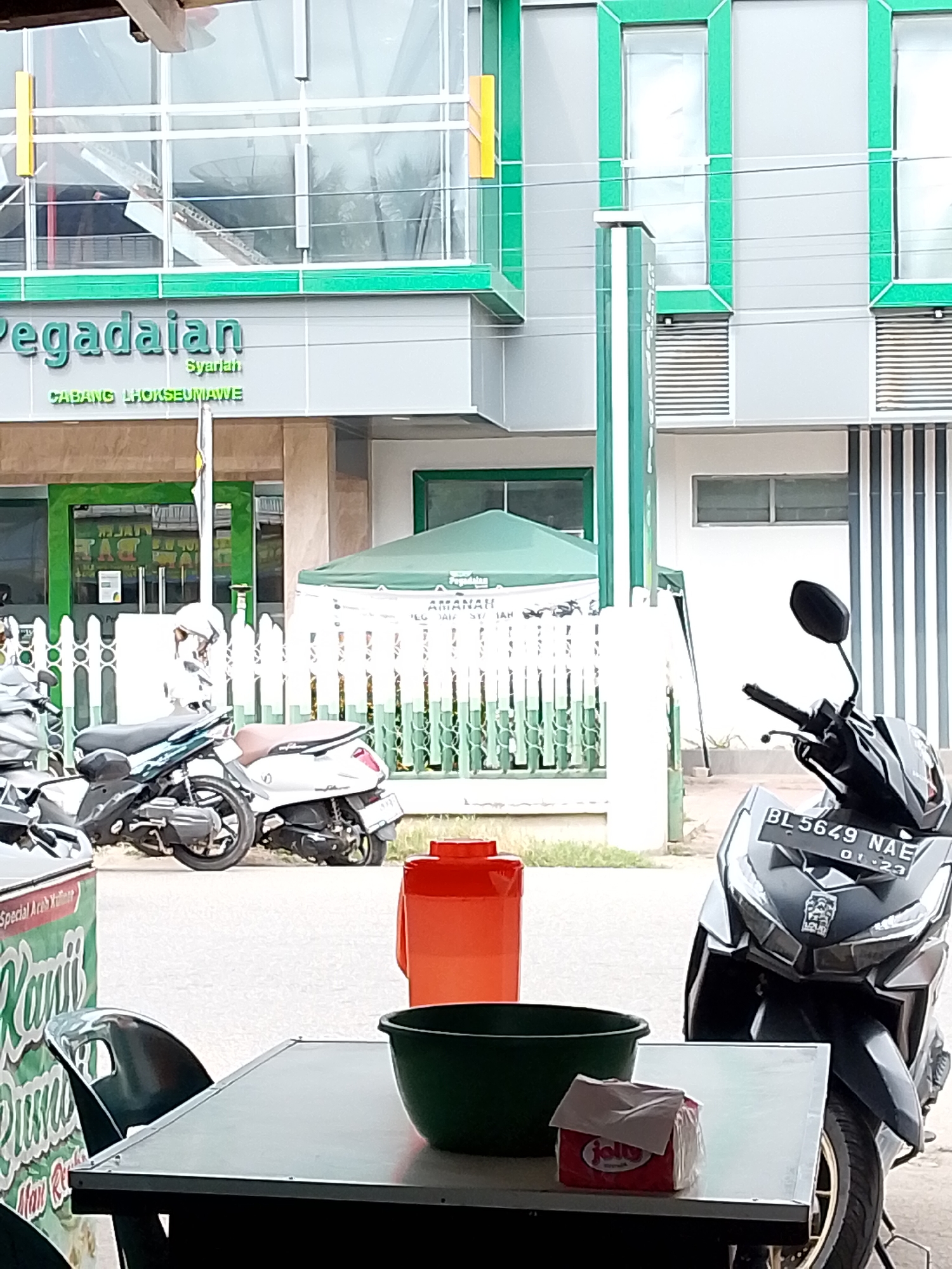 The image depicts a street scene in Lhokseumawe, Indonesia, likely in front of a "Pegadaian Syariah" (Sharia pawnshop) branch. In the foreground, a table is set with a dark green plastic bowl and a package of "Jolly" brand tissues. A dark-colored motorcycle, with license plate "BL 5649 NAE," is parked on the right side of the frame. Behind the table and motorcycles, a white fence with green accents separates the street from the building. The building itself features a prominent "Pegadaian Syariah" sign with "CABANG LHOKSEUMAWE" below it. Green awnings are visible, one extending over the fence and another on the building. Two other scooters are parked near the fence. The overall environment suggests an outdoor setting in a commercial area.